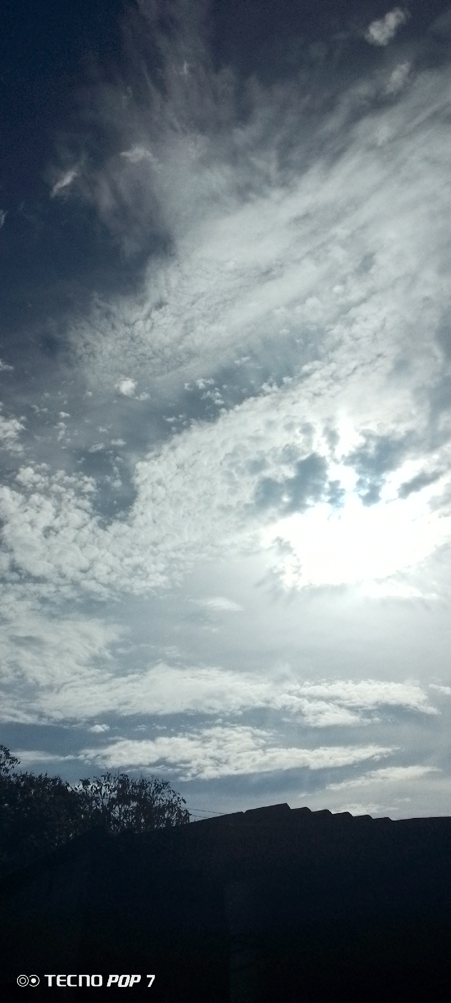 A sky filled with a mix of cirrus and cumulus clouds, the sun shining through, with the silhouette of what appears to be a building and some trees in the foreground. The image was taken with a Tecno Pop 7 phone. There is no information in the image to identify the location as Altagracia, Venezuela, nor the presence of any people, subjects, objects, actions, or interactions beyond what is described above.