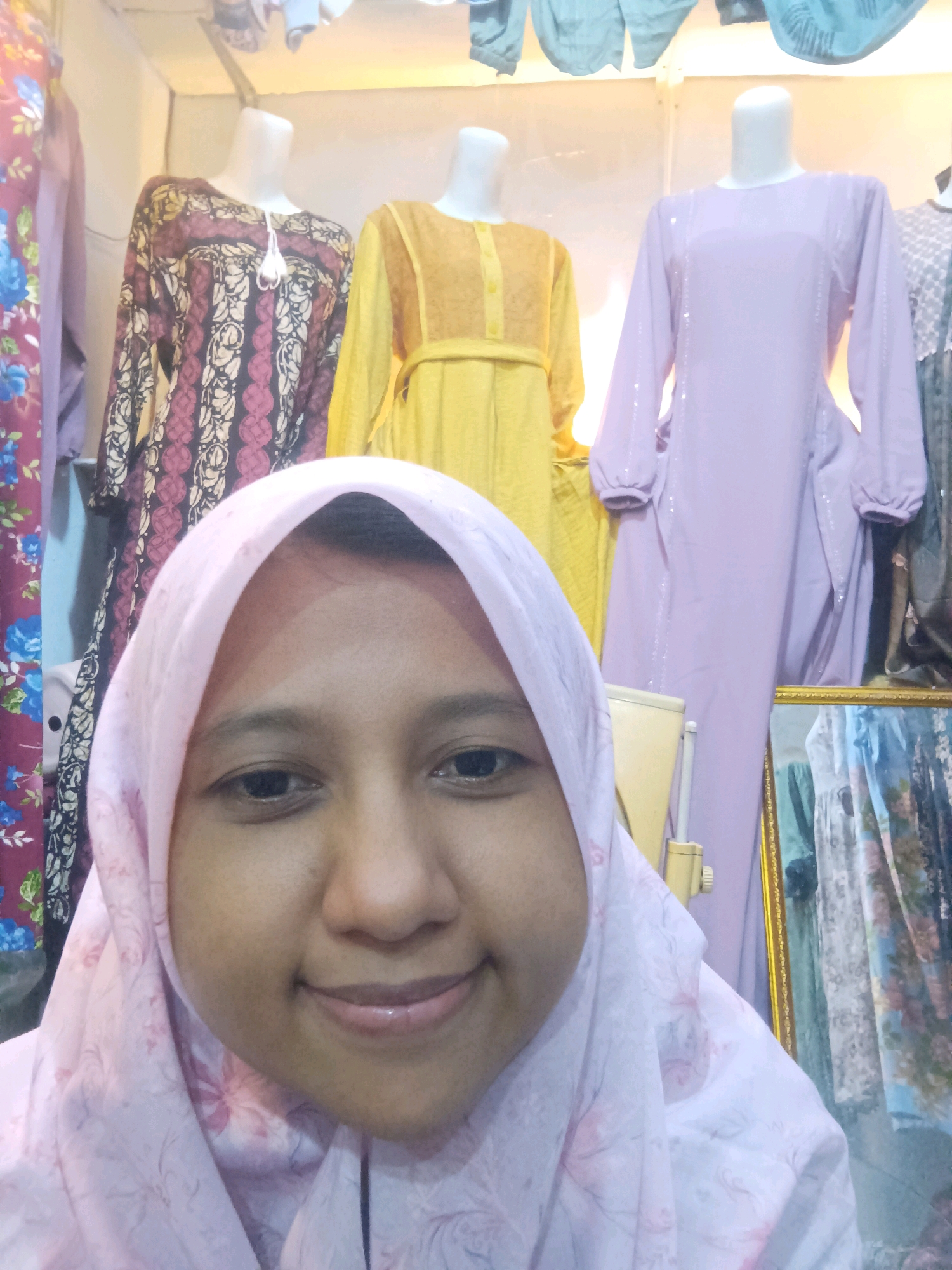 The image shows a woman in a pink hijab taking a selfie in front of mannequins dressed in various garments. The background appears to be a clothing store, with several dresses and fabrics displayed on mannequins and hanging racks. The woman is smiling, and her face takes up a significant portion of the foreground. The store appears to be indoors, with artificial lighting. The location is identified as Sumuragung, Indonesia.