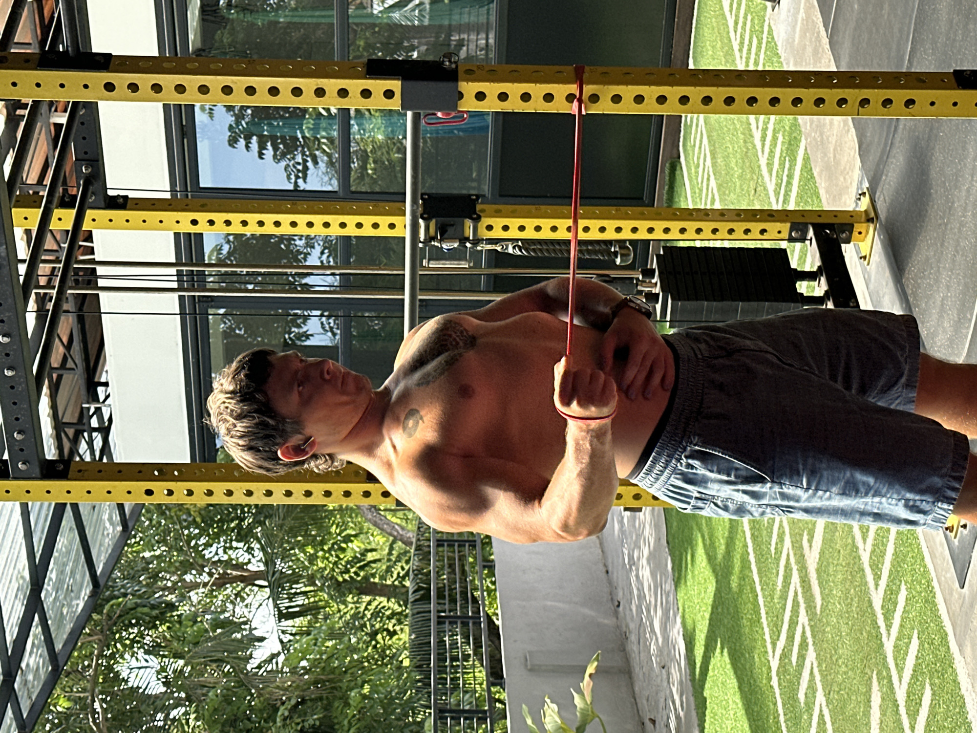 A shirtless man with a tattoo on his chest stands in front of a yellow metal workout rack, pulling on a red resistance band attached to the rack. He is wearing shorts and a watch. The workout rack is situated on a patterned artificial turf surface with white lines, resembling a sports field. Behind the rack, there are windows reflecting trees and other vegetation. The scene is located in North Kuta, Indonesia.