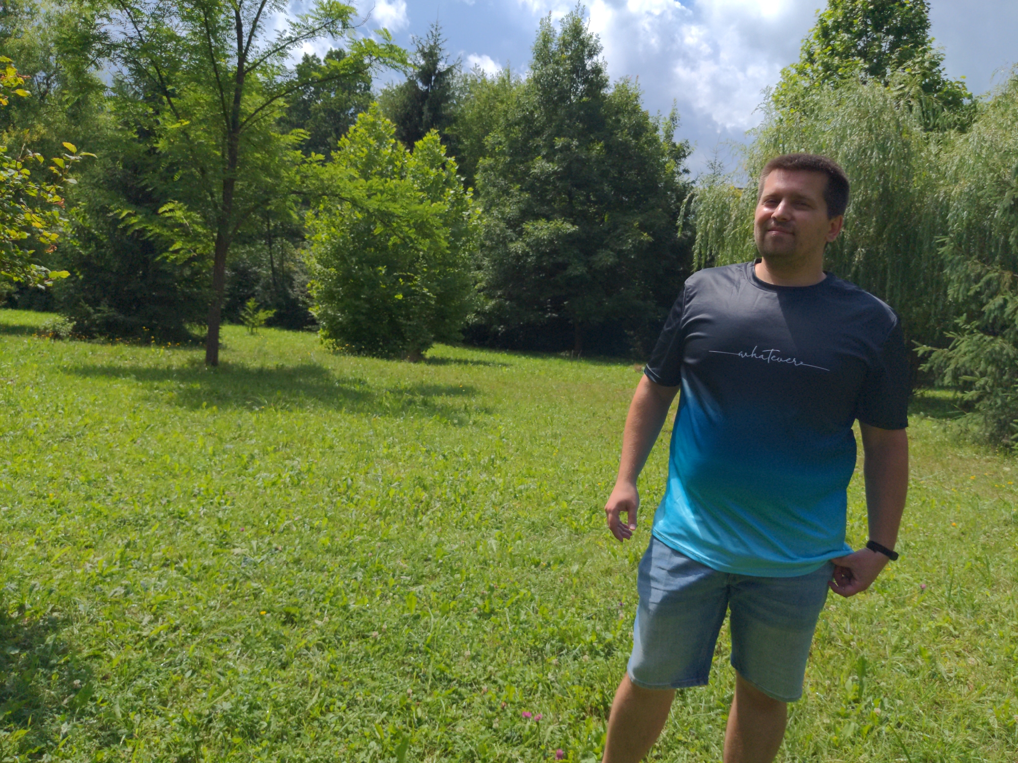In the city of Holubyne, Ukraine, a man stands in a grassy field surrounded by lush green trees under a partially cloudy sky. He is wearing a dark blue t-shirt with a gradient pattern transitioning to light blue and white text that reads "whatever" in cursive, and light wash denim shorts. His right hand is tucked into the waistband of his shorts. The man is looking slightly to his left, with a faint smile on his face. The background features a variety of trees, including some with weeping branches, and the ground is covered in green grass with scattered small wildflowers. Sunlight casts shadows on the grass, indicating it is daytime. The scene is tranquil and appears to be a natural outdoor setting.