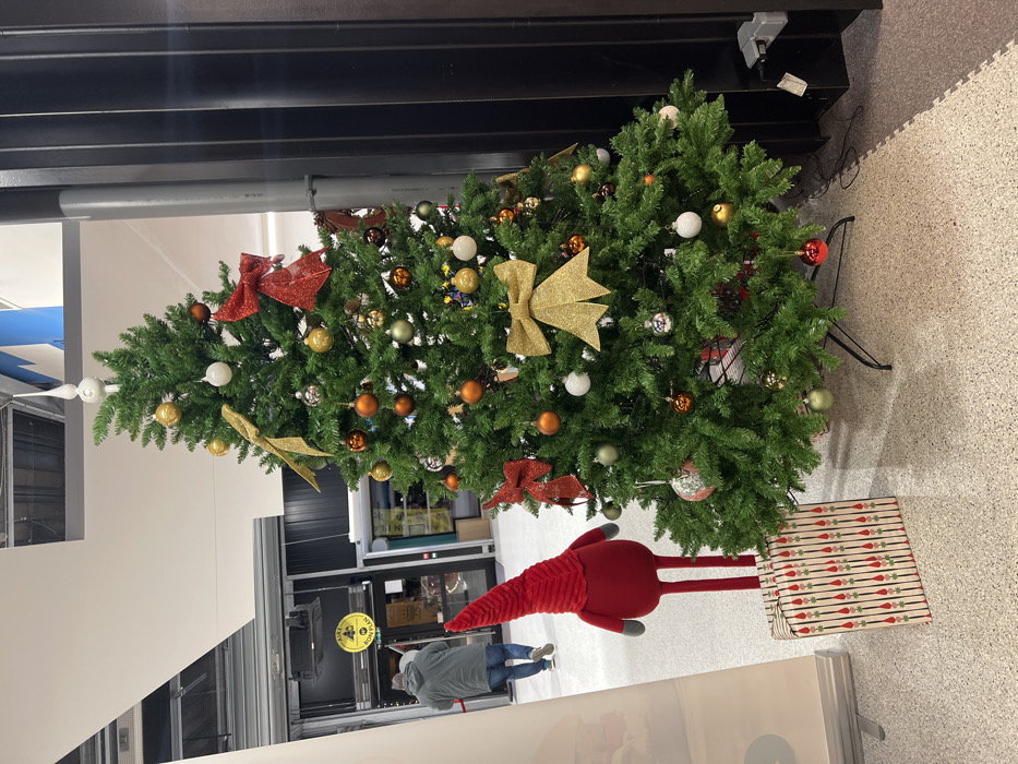 A decorated Christmas tree stands in an indoor space. It's adorned with gold, red, and orange ornaments, bows, and white spherical decorations. A red plush toy with long, thin legs and a striped present are positioned near the base of the tree. A person wearing a grey jacket is visible in the background. The area has a commercial or retail setting.