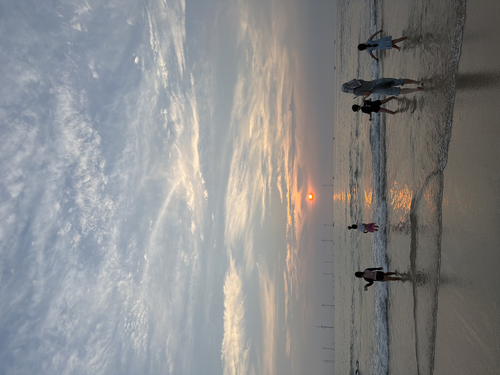 This is a vertical, eye-level, medium shot taken from a beach, showing a group of children and adults wading in the ocean at sunset. The sun, a bright orange disc, is setting on the horizon, casting a warm glow on the water and sky. The sky is filled with wispy clouds in shades of gray, white, and pink, suggesting the last moments of daylight.
In the foreground, the wet sand of the beach meets the gentle waves of the ocean. Several figures are visible in the shallow water. On the left, a child wearing a dark swimsuit and a pink top stands facing away from the camera, holding a phone in their right hand as if taking a picture. To their right, another child in a pink skirt and white top splashes in the water, facing away from the camera. In the center right, an adult wearing a headscarf and light-colored pants stands with their back to the camera, facing the sea. To the adult's left, a child in a dark t-shirt with a number "7" on it stands facing the sea. To the adult's right, a child in a light blue dress faces away from the camera.
In the distance, a line of wind turbines stands tall in the sea, silhouetted against the sunset. Several small boats are also visible on the water. The scene suggests a peaceful and recreational atmosphere. Given the presence of wind turbines in the ocean and the characteristic sunset over the water, this scene is likely from the coast of Jeju Island, South Korea, possibly near an offshore wind farm.