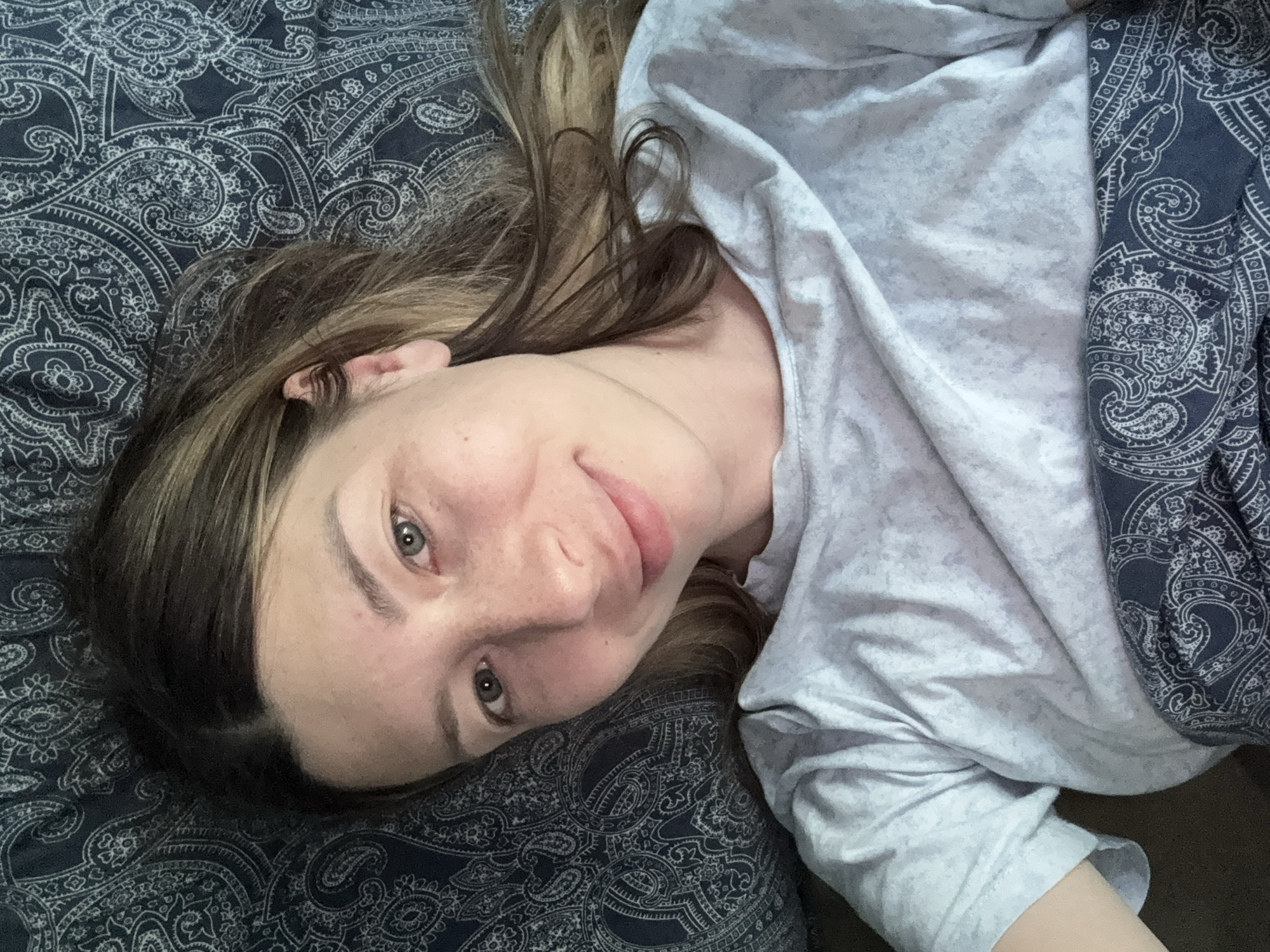 The image is a selfie taken from a high angle, looking down at a young woman lying in bed. She has light brown hair with blonde highlights, fair skin, and green eyes. She is wearing a light grey t-shirt. She is lying on a bed with dark blue patterned sheets. The image is taken indoors, in what appears to be a bedroom. There is no information in the image to suggest the location is in Moscow, Russia.