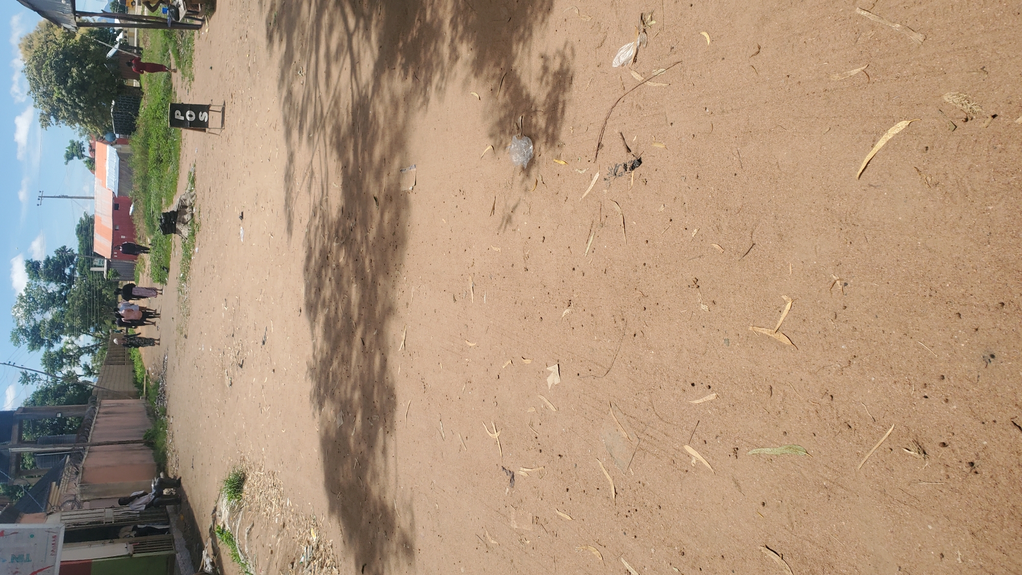 The image depicts a dirt road in the city of Jalingo, Nigeria. On the left side of the road, a building with a metal gate and some construction is visible, with a person sitting on a raised platform outside. Further down the road, a group of individuals, appearing to be locals, are walking away from the camera. One person is wearing a dark suit, while others are in casual attire. A red and orange structure, possibly a small building or a stall, is situated in the background. To the right, a small sign with "POS" written on it in black lettering is propped up on the ground. The road is unpaved and dusty, with scattered leaves and debris. The environment appears to be a semi-urban or rural setting, with trees and a partly cloudy sky overhead.
