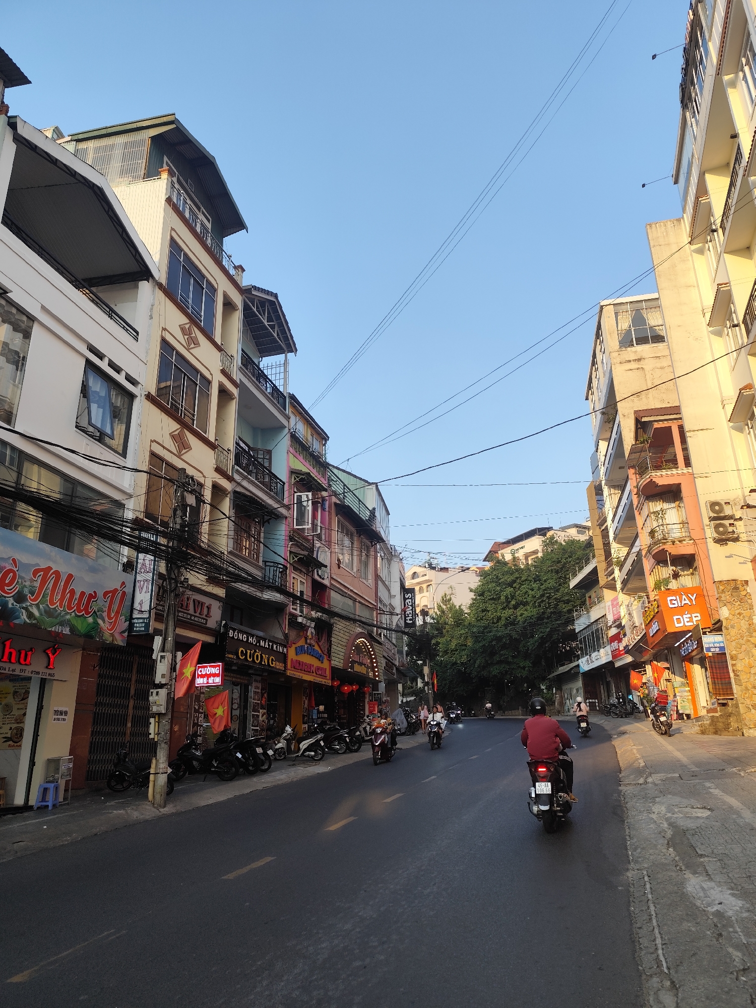 The image depicts a street scene in Da Lat, Vietnam. The view is from a low angle on the street, looking towards a slight incline. The street is asphalt and has a few motorcycles driving on it. Parked motorcycles line the left side of the street. The buildings are multi-story and appear to be a mix of residential and commercial, with various signs in Vietnamese displayed. Some businesses have red banners with yellow stars, likely Vietnamese flags. Power lines crisscross above the street. Trees are visible further down the street. The sky is blue and the lighting suggests it is either early morning or late afternoon.