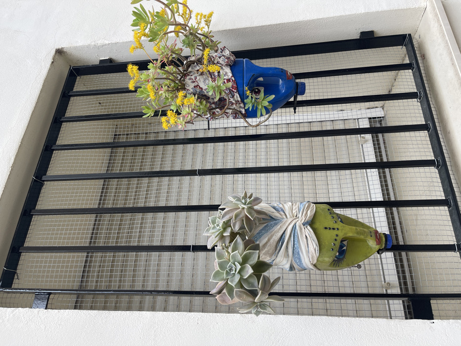 The image displays an exterior view of a building in Buñol, Spain, centered on a window recessed into a white, textured plaster wall. A black metal security grate, consisting of seven visible vertical bars and horizontal top and bottom supports, covers the window opening. A fine, light-colored square mesh screen is affixed to the interior side of the metal grate. Behind the grate, a light-toned roller shutter (persiana) with horizontal slats is visible.
Two decorative planters, fashioned from repurposed plastic containers and stylized as faces, are suspended from the black grate by wire hooks. The planter on the left is a light green container featuring blue painted eyes, black eyeliner, red lips, and multiple silver studs on its 'face.' A beige and light blue striped fabric is tied around the top, resembling a turban. This planter holds several succulent plants characterized by fleshy, greenish-grey rosettes. The planter on the right is a blue container with white painted eyes, black pupils, a small red mouth, and a small green plant emerging from its side. A colorful fabric with a dark background and red, white, and blue patterns is wrapped around its upper portion. This planter contains a plant with numerous small, bright yellow flowers and green, fleshy leaves. Natural daylight illuminates the scene.