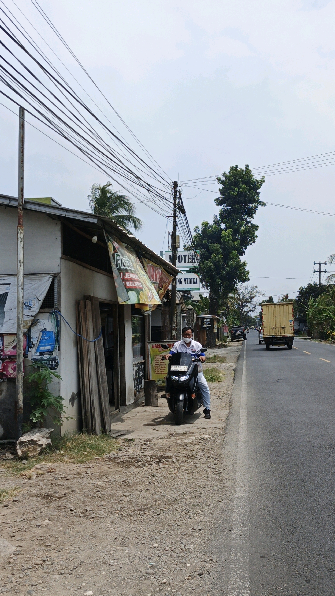 A man wearing a white shirt with blue accents and a white face mask is sitting on a black scooter on the side of a road in Desa Wonoharjo, Indonesia. The scooter is parked on a gravelly patch next to a small, weathered building with a green awning. The building appears to be a shop or a small business, with various signs and posters displayed on its exterior. To the right of the building, a large green tree stands tall. Across the paved road, a yellow truck is traveling away from the camera. The sky is overcast.
