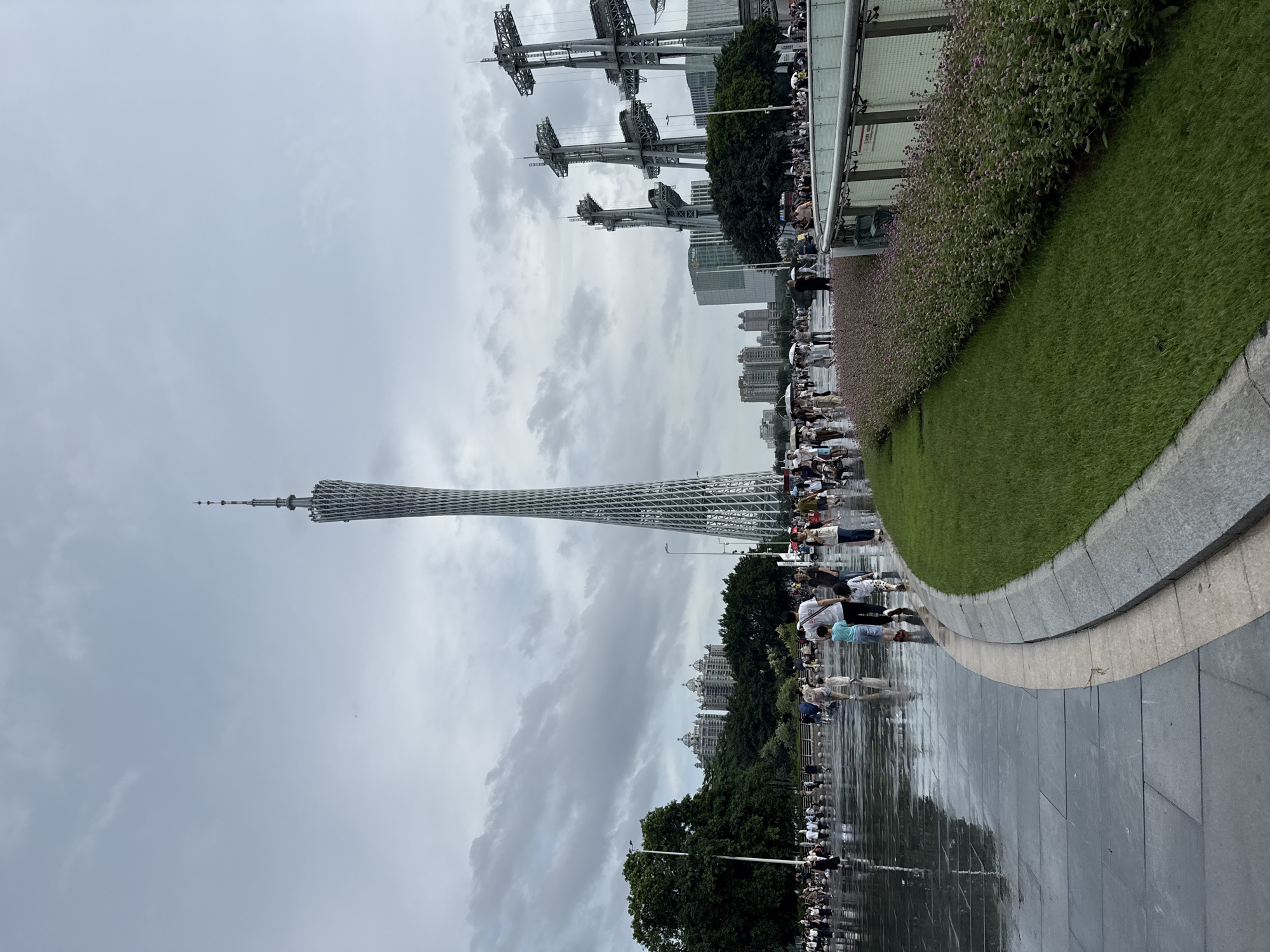 The image depicts a scene in Tianhe District, China, with the Canton Tower prominently featured as a central landmark. The weather appears overcast, with a cloudy sky. The foreground shows a wet, paved plaza where numerous people are walking and strolling. The ground reflects the surroundings, indicating recent rain or dampness. To the right of the plaza, a well-maintained strip of green grass and flowering plants borders a curved walkway. In the mid-ground, beyond the plaza, are more people, trees, and several other modern buildings and structures, including what appear to be large, industrial-looking towers with intricate metalwork. The Canton Tower, a tall, slender, lattice-like structure, rises majestically in the background, dominating the skyline. The overall atmosphere is one of a public urban space, possibly a park or a waterfront area, bustling with pedestrian activity under a subdued sky.
