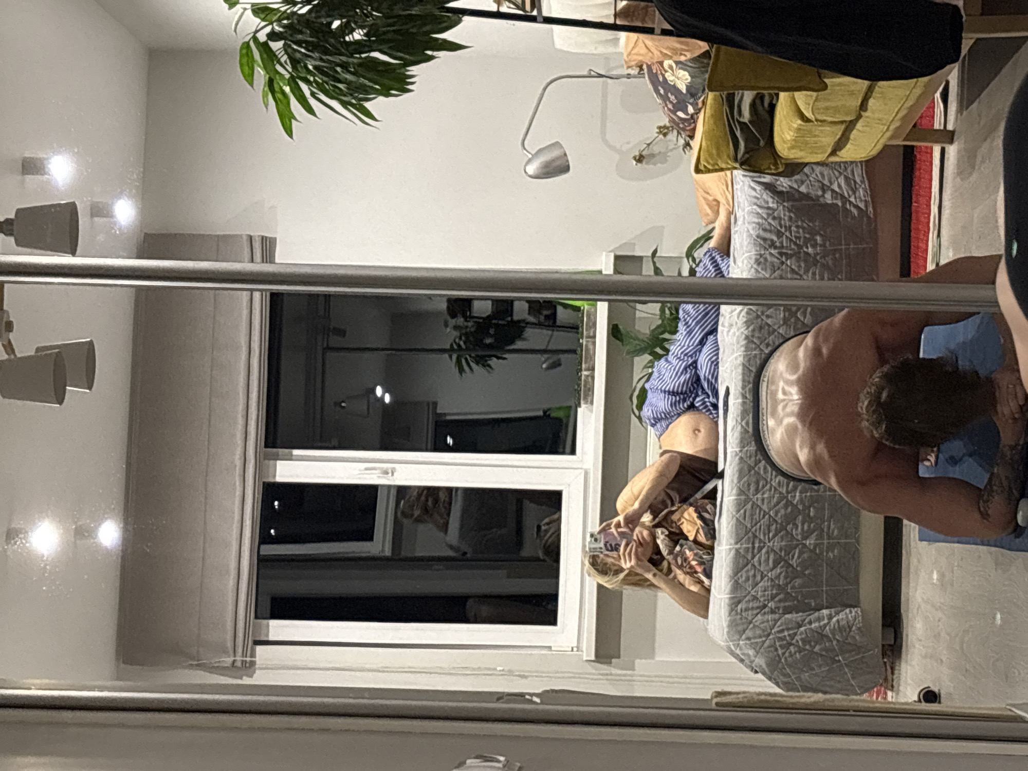 The media file depicts an indoor scene, likely a living room, with a focus on two individuals. One person, with their back to the camera and appearing shirtless, is positioned on a light grey quilted surface that resembles a massage table or bed. Their musculature is visible, and they are holding a blue object in their hands. Another person is captured from the side, seated and holding a smartphone, seemingly taking a picture. This individual is wearing a patterned garment with floral motifs and blue striped pants. The room features a large window with blinds, through which the exterior, possibly a nighttime scene, is visible, showing lights. A tall green plant is situated in the upper left corner. The lighting in the room appears to be artificial, with spotlights on the wall. The overall impression is that of a private, relaxed setting. There is no visible text in the media. The location context is Nizhny Novgorod, Russia.