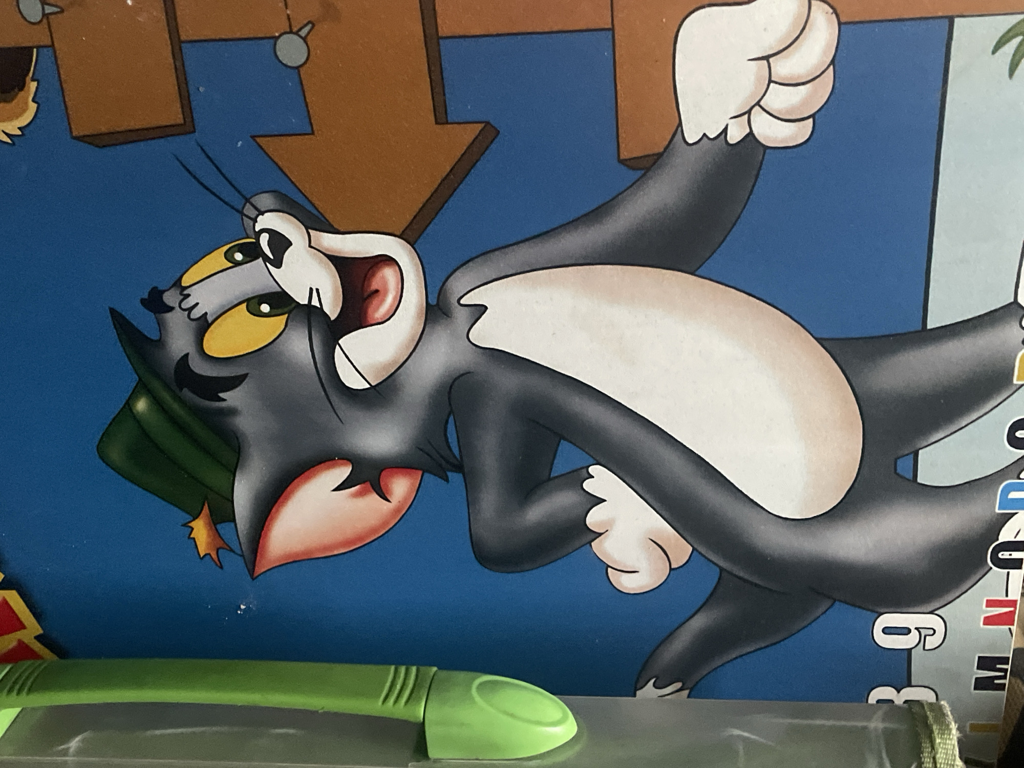 The image shows a close-up of a cartoon depiction of Tom, a cat character from the Tom and Jerry series, with a green marker pen in the foreground. Tom is in a dynamic pose, seemingly suspended in mid-air, with his mouth open and arms outstretched. He has a green object (possibly a hat or leaf) on his head. The background is a blue gradient with a brown arrow pointing downwards. The image includes part of the text "DUN 6".
I am unable to provide information about the location in Dhaka, Bangladesh as it is not discernible from the image.
