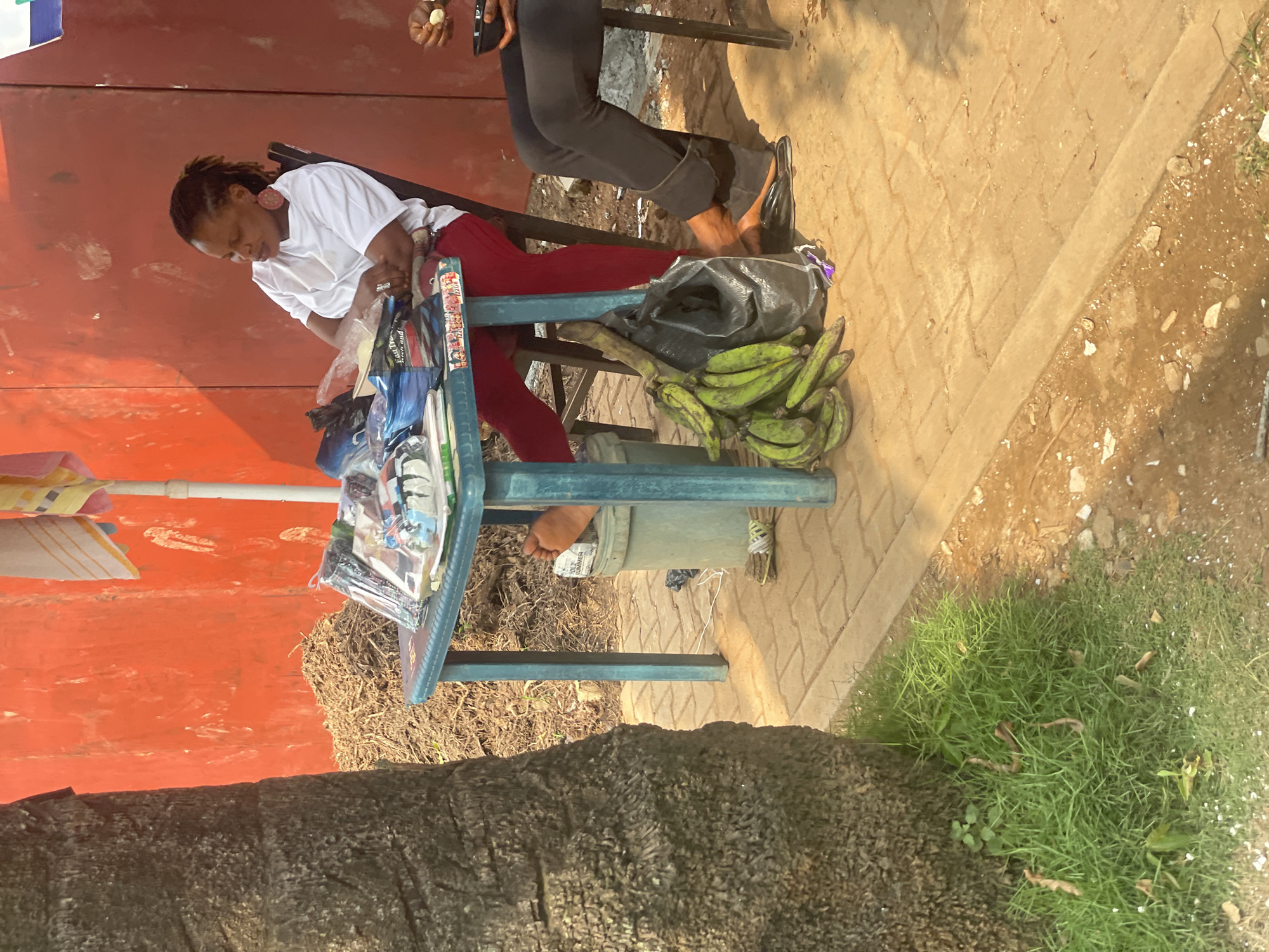In Abuja, Nigeria, a woman sits at a turquoise plastic table laden with merchandise, including packaged clothing and accessories. She is wearing a white t-shirt, red trousers, and large hoop earrings. Her feet are bare, and one is resting on a grey bucket. A bunch of green plantains is in a black plastic bag on the ground next to her. The scene is set against a vibrant orange wall, with a palm tree trunk to the left and dry brush behind the table. Another person's legs are visible to the right, seated on a black chair, holding a bowl. The ground is paved with interlocking bricks and some sandy soil.