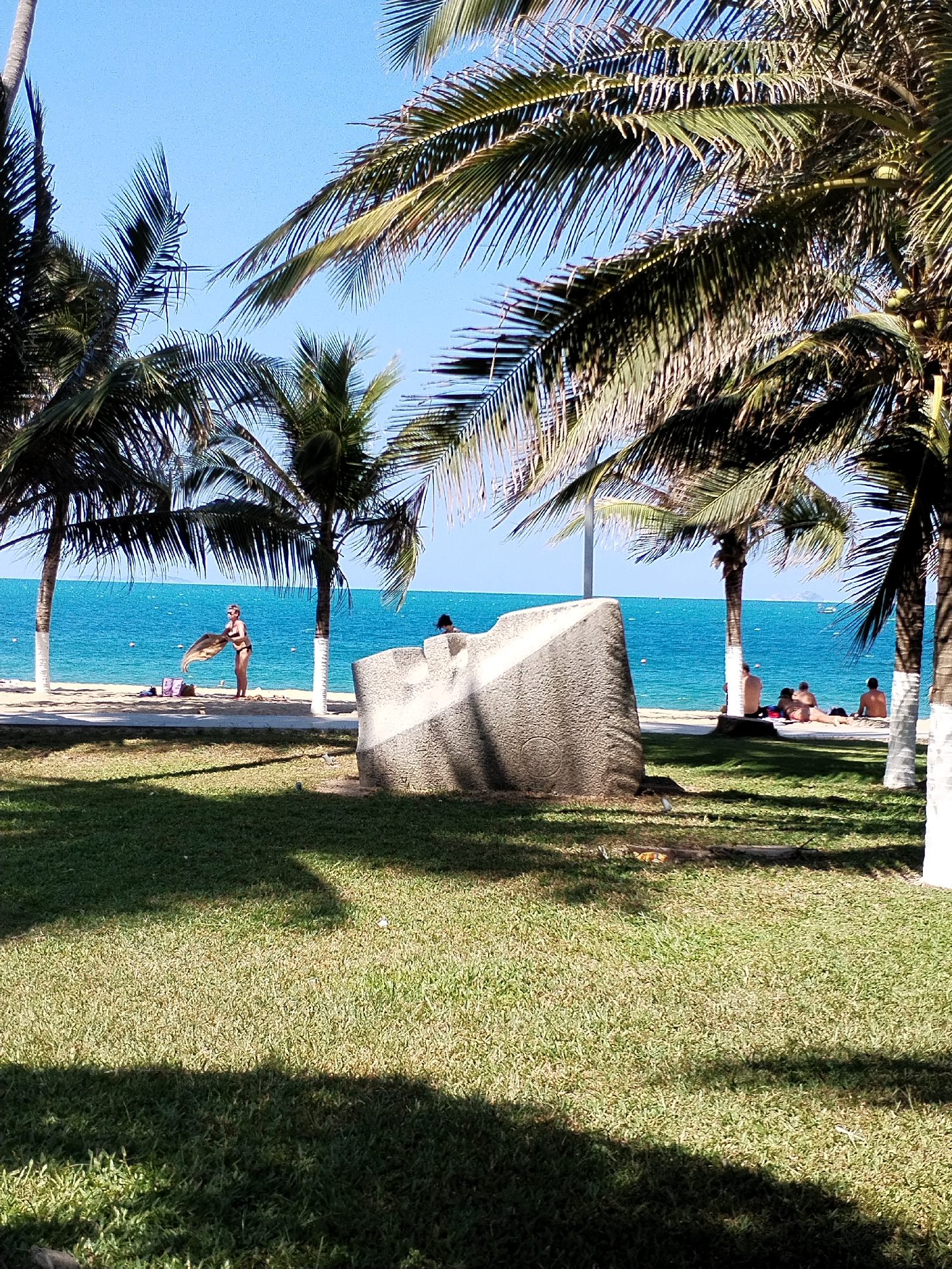 The image shows a tropical scene in Nha Trang, Vietnam. The foreground is a grassy area with shadows from palm trees. A large, grey, roughly triangular concrete structure stands between the grass and a paved walkway. Beyond the walkway is a beach with light sand and blue water. Several people are on the beach, including a woman holding a towel near a beach bag and a group of people sunbathing under a palm tree with a whitewashed trunk. Other palm trees are scattered throughout the scene. The sky is clear and blue.