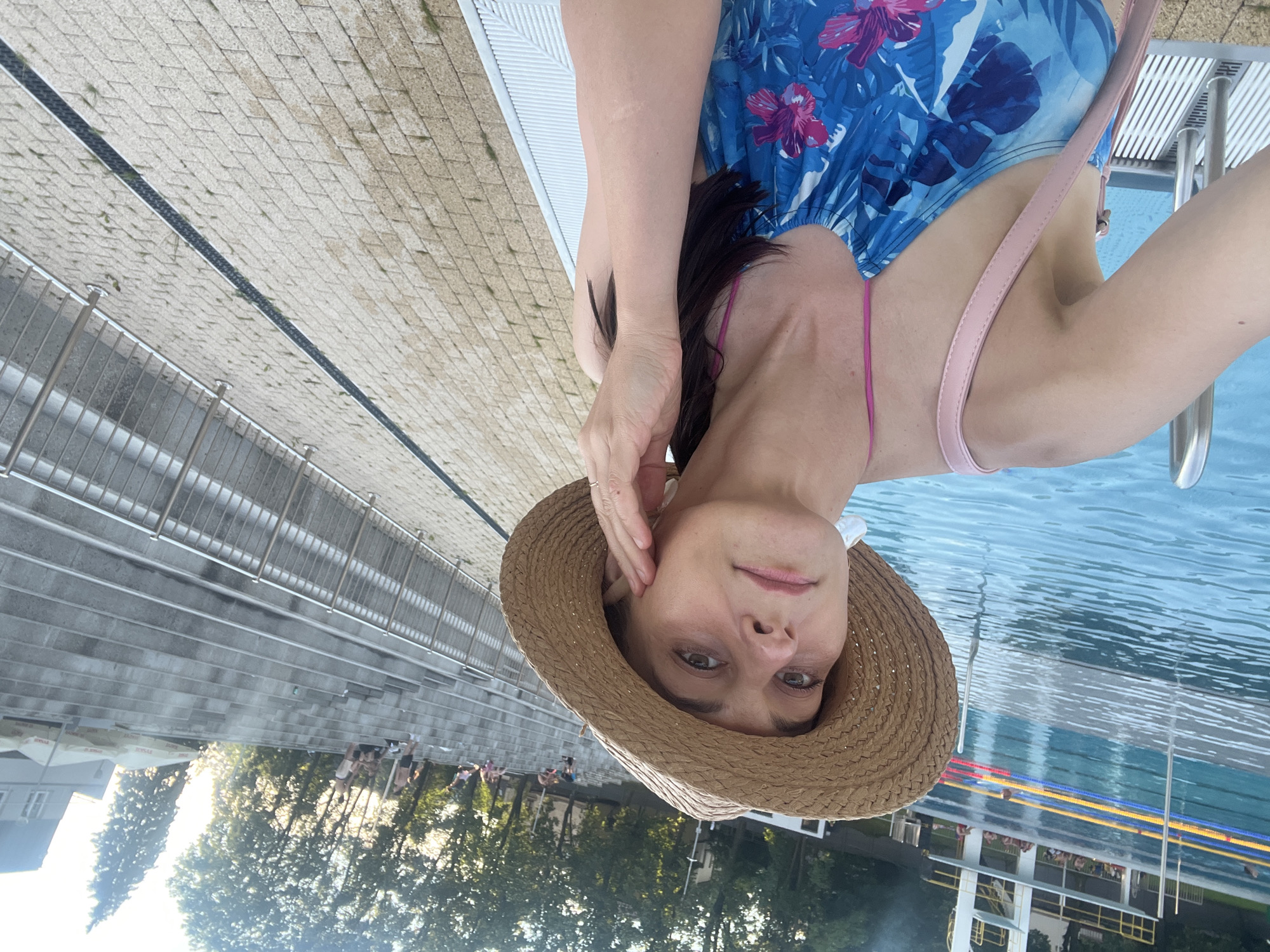 A woman wearing a straw hat and a blue floral swimsuit poses for a selfie by a swimming pool. The pool has lanes marked with blue, yellow, and red lines, and a diving platform is visible in the background. To the right of the pool, there are tiered concrete steps with people sitting on them. A pink strap from the woman's swimsuit is visible, as is a pink purse slung over her shoulder. The ground around the pool is paved with light-colored bricks. The lighting suggests it is late afternoon or early evening.
