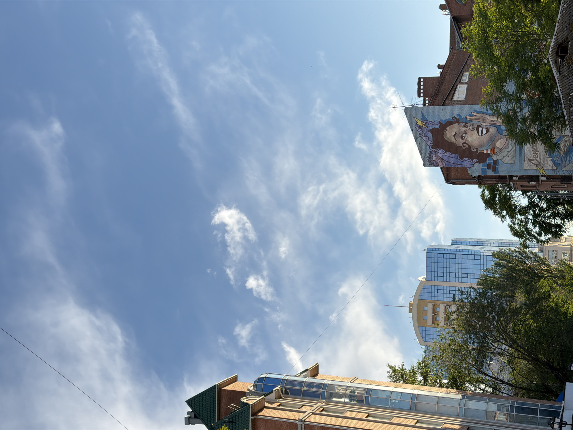 The image shows a view of the sky above the city of Rostov-on-Don, Russia, framed by buildings. In the top right, a brick building has a large mural of a woman on its side, partially obscured by trees. Below that, a modern glass-covered building rises, also partly hidden by trees. At the bottom of the image, another brick building with a green roof is visible. Overhead electrical lines crisscross the sky. The sky is blue with scattered wispy clouds.