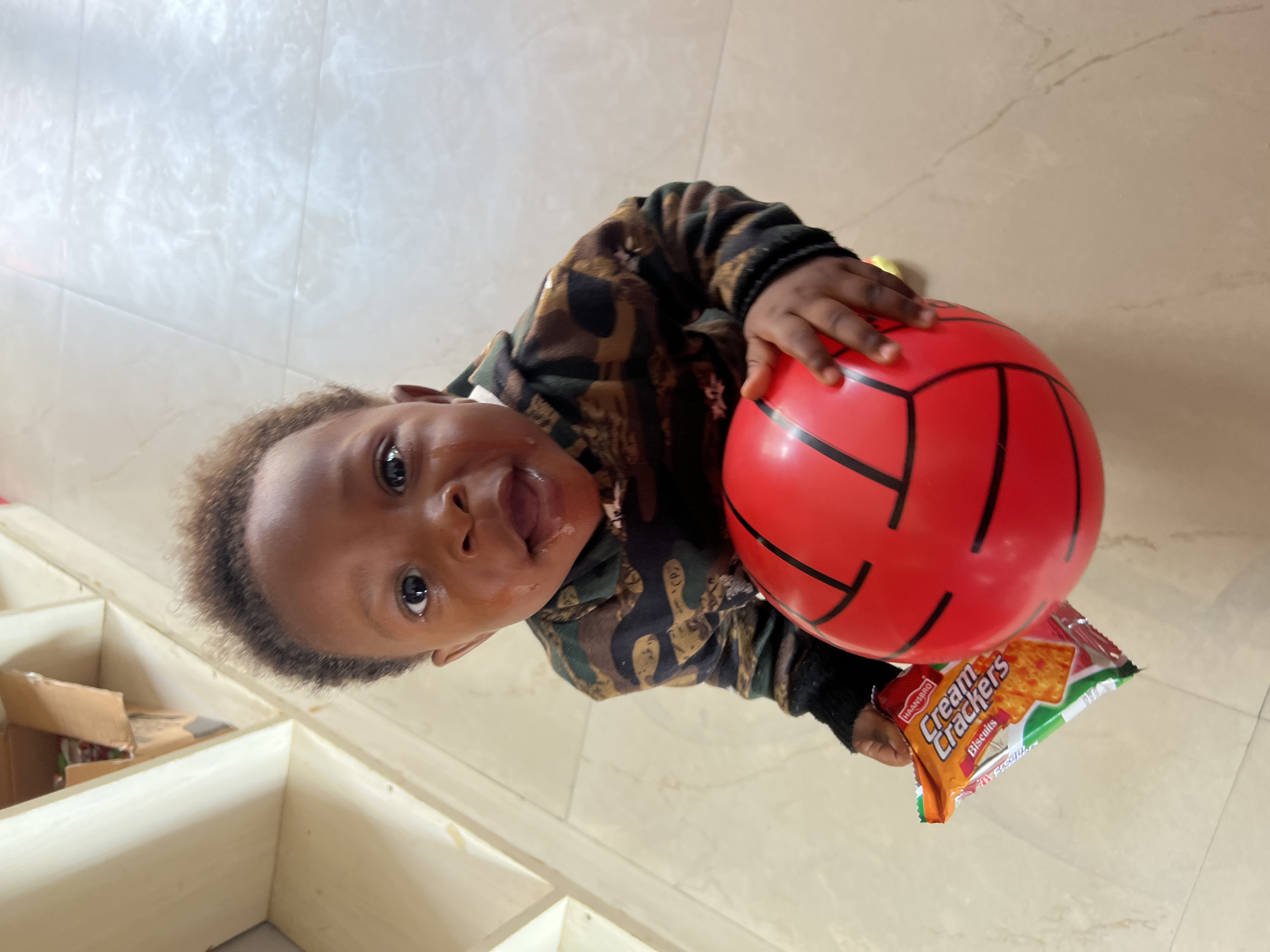 A toddler stands in a room with light-colored tile flooring. The child, wearing a camo-patterned top, holds a red ball in one hand and a packet of "Cream Crackers" biscuits in the other. To the side of the toddler, there is a white shelving unit with some cardboard boxes inside. The location is Gura Top, Nigeria.