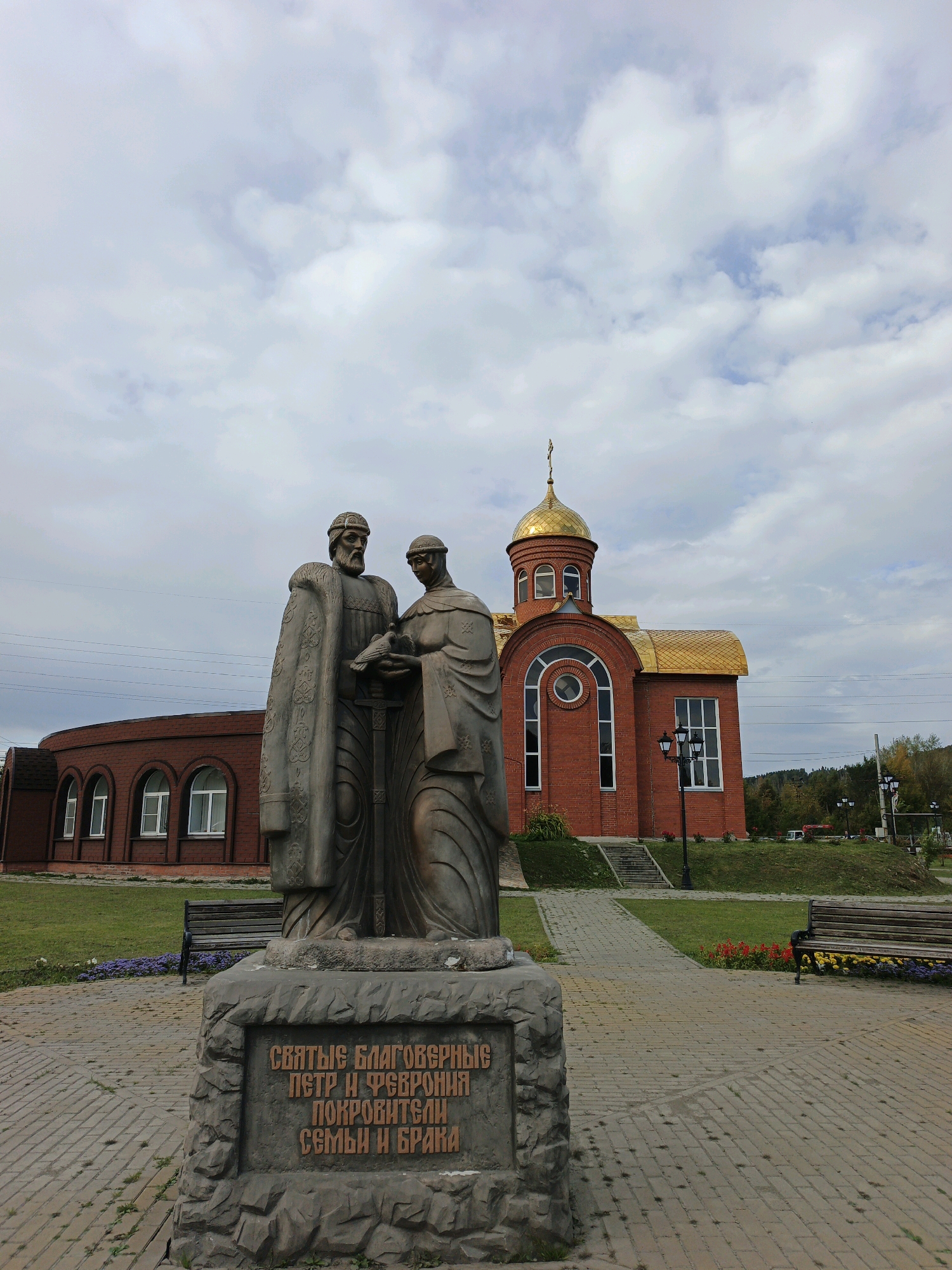 In the city of Zlatoust, Russia, a statue of Saints Peter and Fevronia stands in the foreground, positioned on a stone pedestal inscribed with Russian text. The statue depicts a man and a woman, both dressed in historical attire, with the man holding a dove. Behind the statue, a brick building with a golden dome, resembling a church, is visible. The building features arched windows and is surrounded by a paved walkway, grassy areas, and benches. The sky above is cloudy.