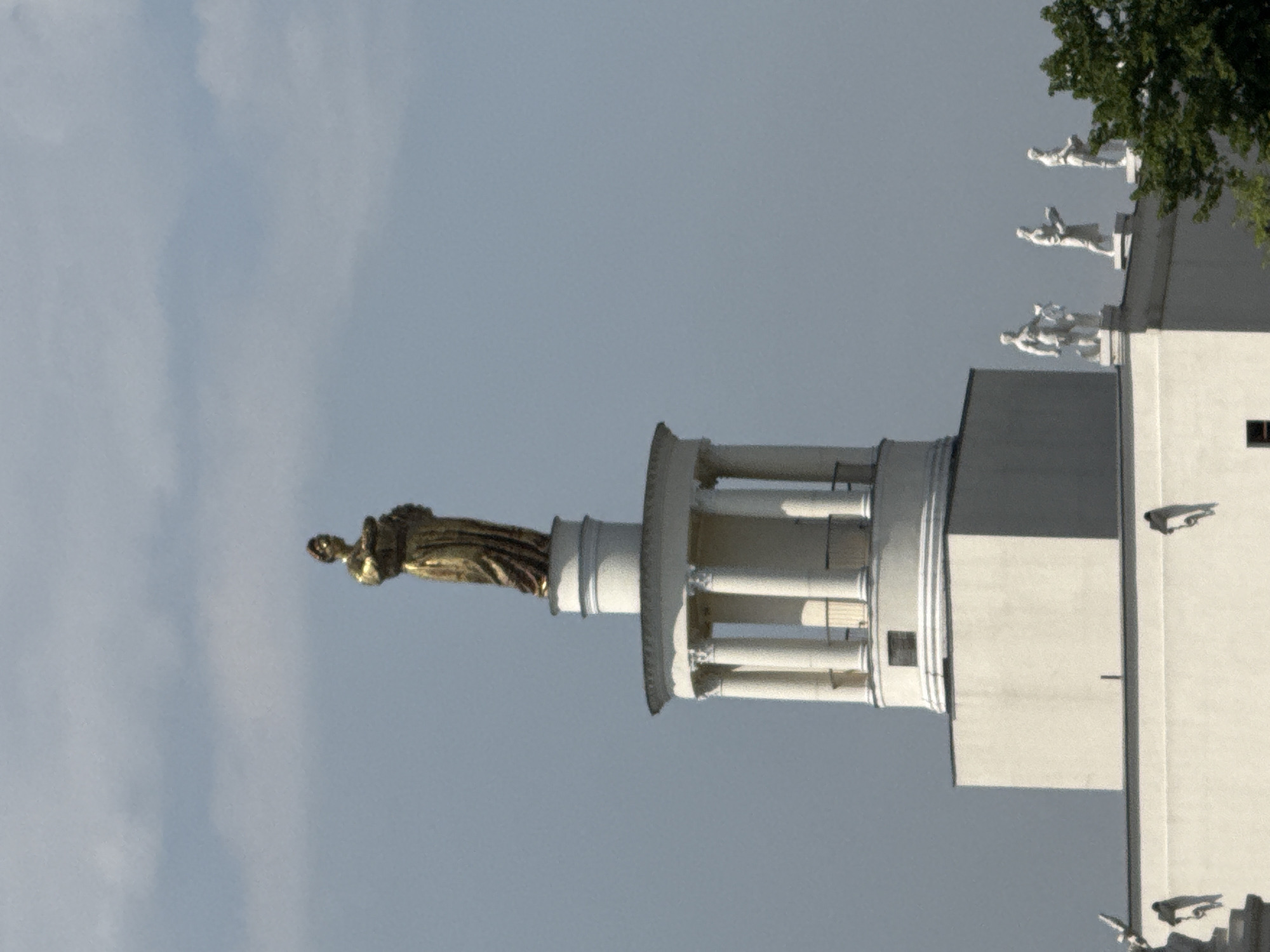 A low-angle shot depicts the Central Pavilion of the VDNKh exhibition park in Moscow, Russia, showcasing the golden statue "Worker and Kolkhoz Woman" against a soft gray sky. The architectural details of the pavilion, including its columns and sculptures on the rooftop, are clearly visible. The image captures a portion of the building's side facade as well. The environment is bright, suggesting daylight.
