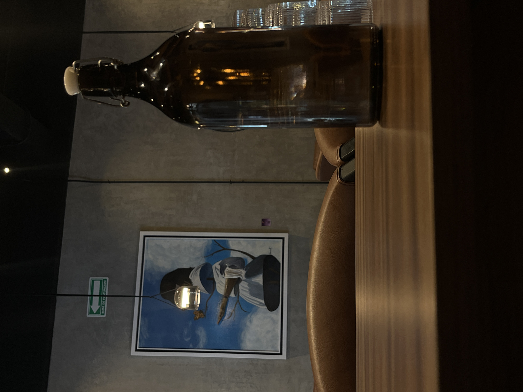A horizontally-oriented image captures a brown glass bottle with a white stopper, sitting on a wooden table. Several clear glasses are stacked behind the bottle. A brown leather chair is adjacent to the table. On the wall in the background, there is a framed painting featuring a blue and white design and a green and white emergency exit sign. The location appears to be an interior space, possibly in an urban area, but the specific city and location are unclear.