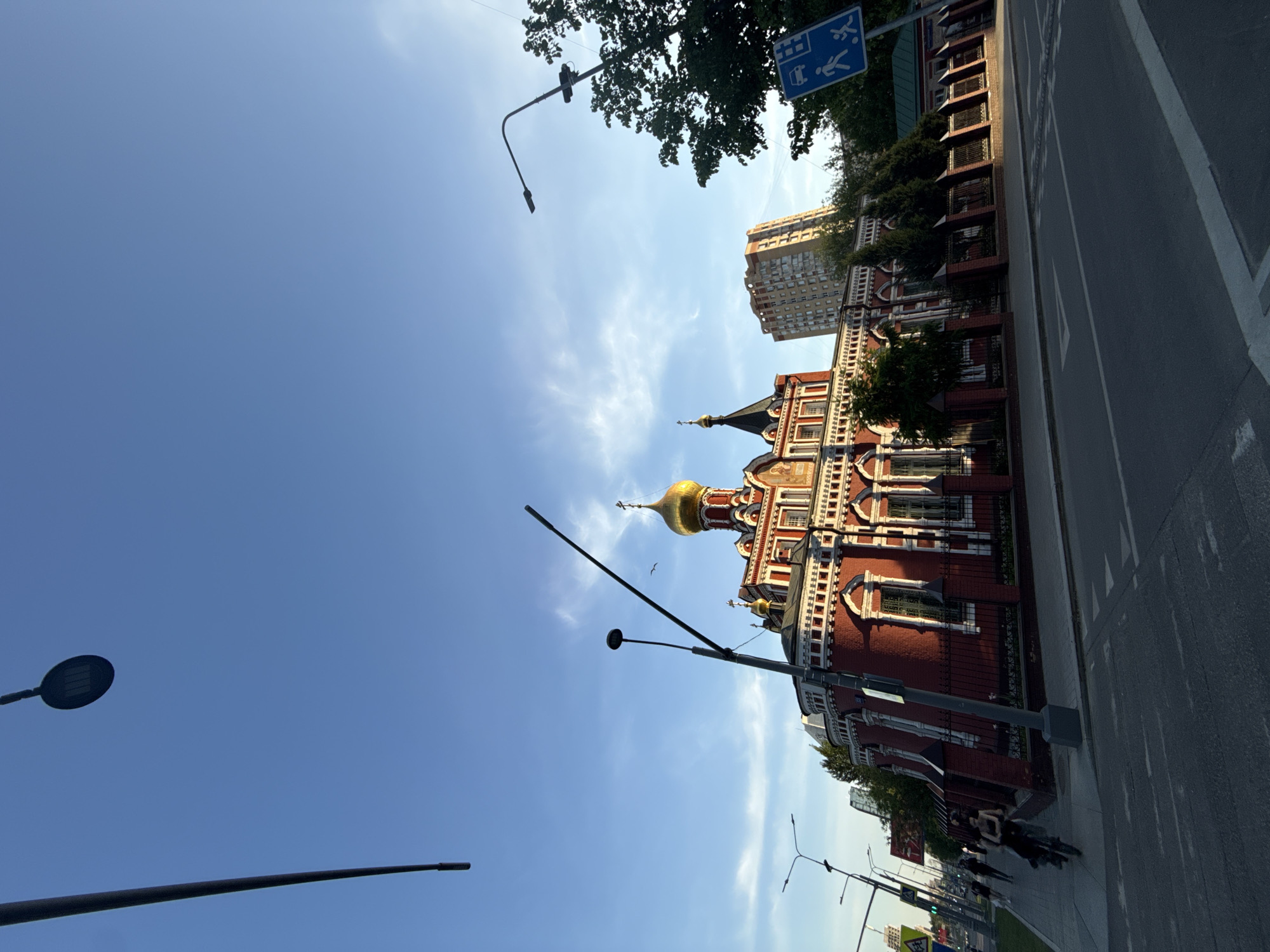 The image depicts a view of the Holy Trinity Church in Moscow, Russia, taken from street level. The church is a red brick building with white decorative elements and golden domes. A modern residential building is located directly behind the church. In the foreground of the image, there is a street with sidewalks and traffic markings. Several lampposts are visible in the image against a blue sky with wispy clouds. The presence of a traffic sign indicates the area is accessible for pedestrians with disabilities. Some people are walking on the sidewalk next to the road.