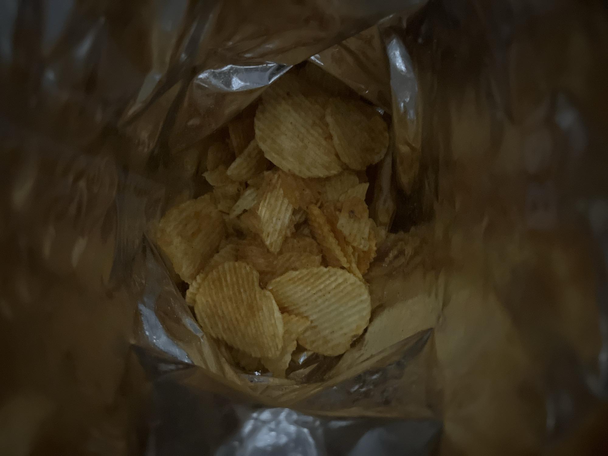 A close-up, overhead shot shows ruffled potato chips inside a shiny, crinkled bag. The chips are golden brown and have a textured, ridged surface. The bag's interior is reflective, showing some blurred highlights and shadows. The scene is likely indoors, with the lighting casting a warm, somewhat dim glow on the chips. There is no information about the location in Vratsa, Bulgaria.