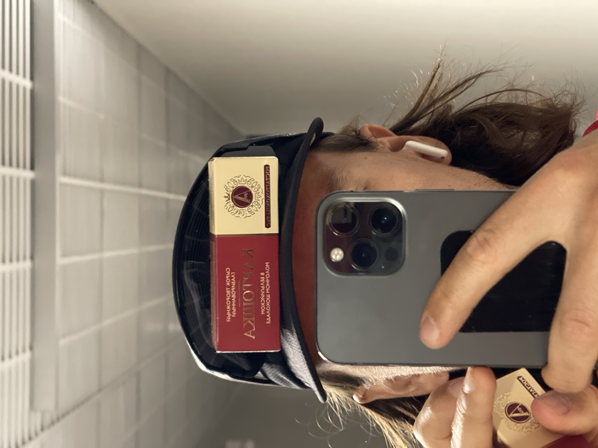 A person wearing a black hat is taking a selfie in a bathroom. The person is holding a gray smartphone in their right hand. On the brim of the hat sits a small, red and gold box of matches. An earbud is visible in the person's right ear. The background shows white tiled walls. The location is Unknown, Unknown.