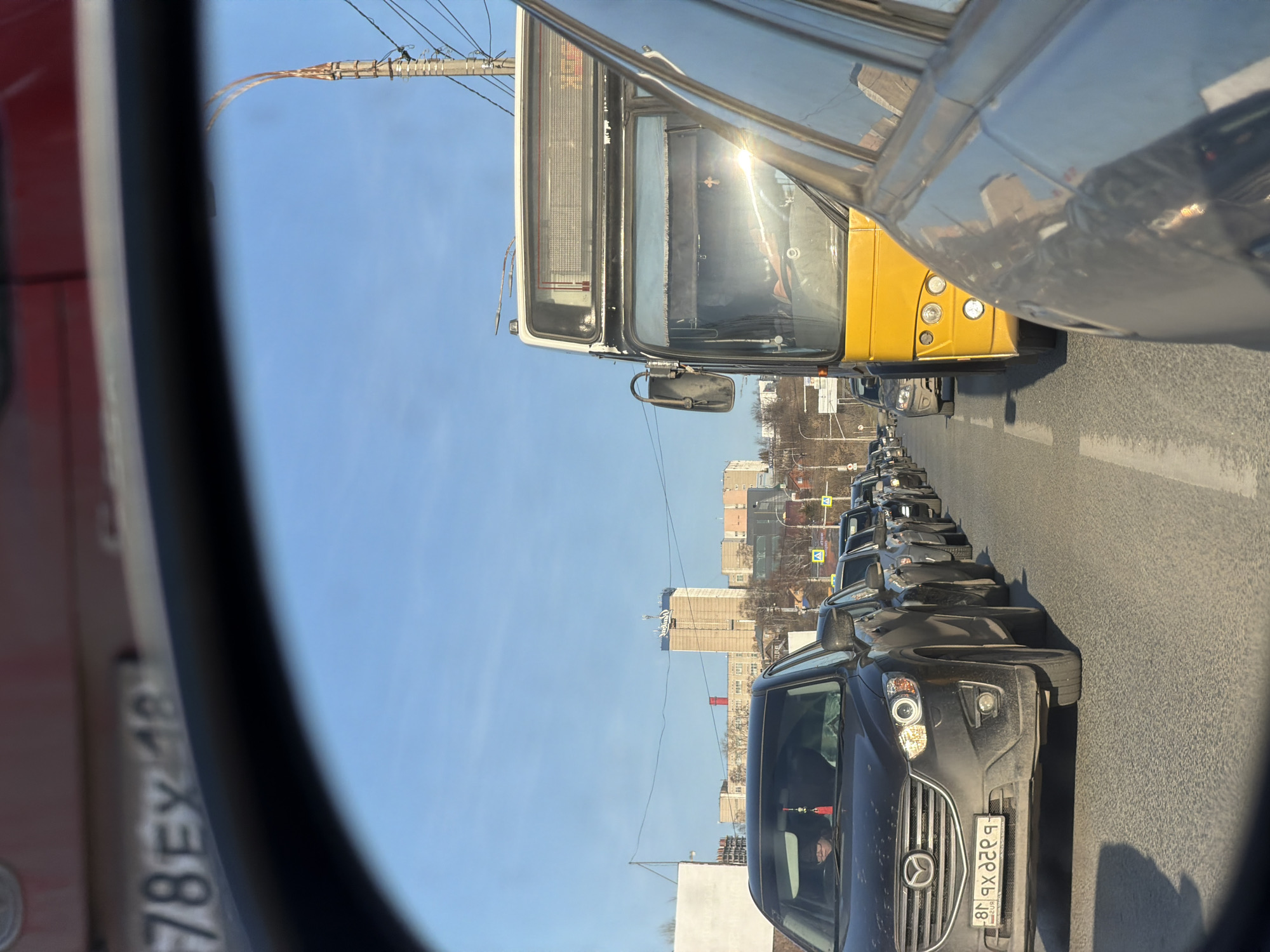 The image, taken from a side mirror of a vehicle, shows a traffic jam on a sunny day in Izhevsk, Russia. A yellow bus is visible in the adjacent lane. Several dark-colored passenger vehicles are lined up in the traffic, with a black Mazda SUV in the foreground. Buildings are visible in the background, with one taller building displaying a "Coca-Cola" sign on top. Power lines stretch across the sky. The number plate of the Mazda is visible as "Я922 XP 18".