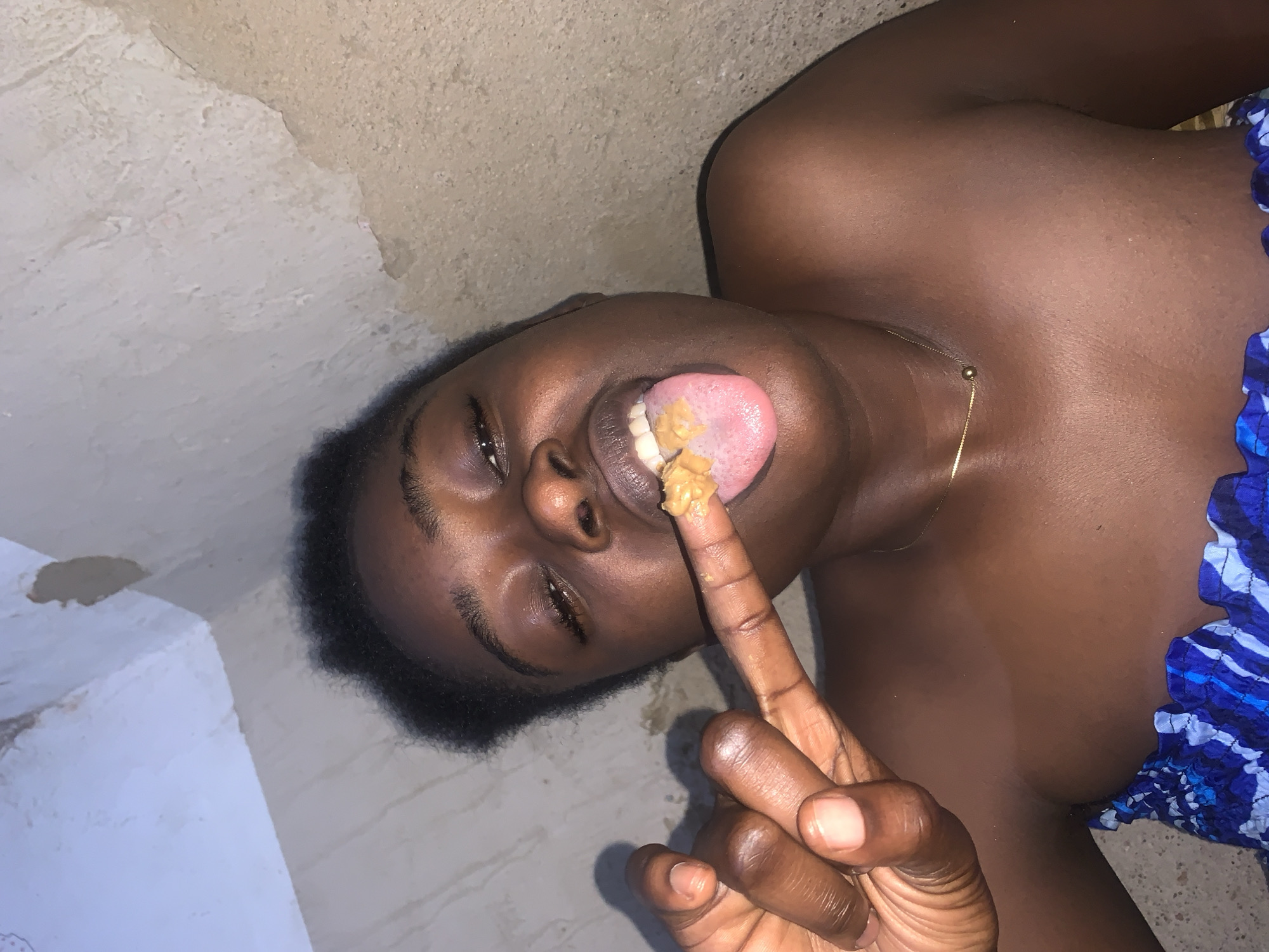 A young Black woman is captured in a close-up shot, her face angled towards the camera with her eyes partially closed. She is holding her right index finger up, with a small amount of a tan substance, possibly peanut butter, on the tip. Her tongue is extended, reaching towards the substance on her finger. The background appears to be an interior space with textured, light-colored walls, exhibiting some peeling paint or plaster in the upper left quadrant. The woman is wearing a blue and white patterned garment that appears to be strapless, and a thin gold chain with a small ball pendant is visible around her neck. The lighting suggests an indoor setting, possibly at night or in a dimly lit room. The image does not provide any specific indicators of its location within Rafin Par, Nigeria.