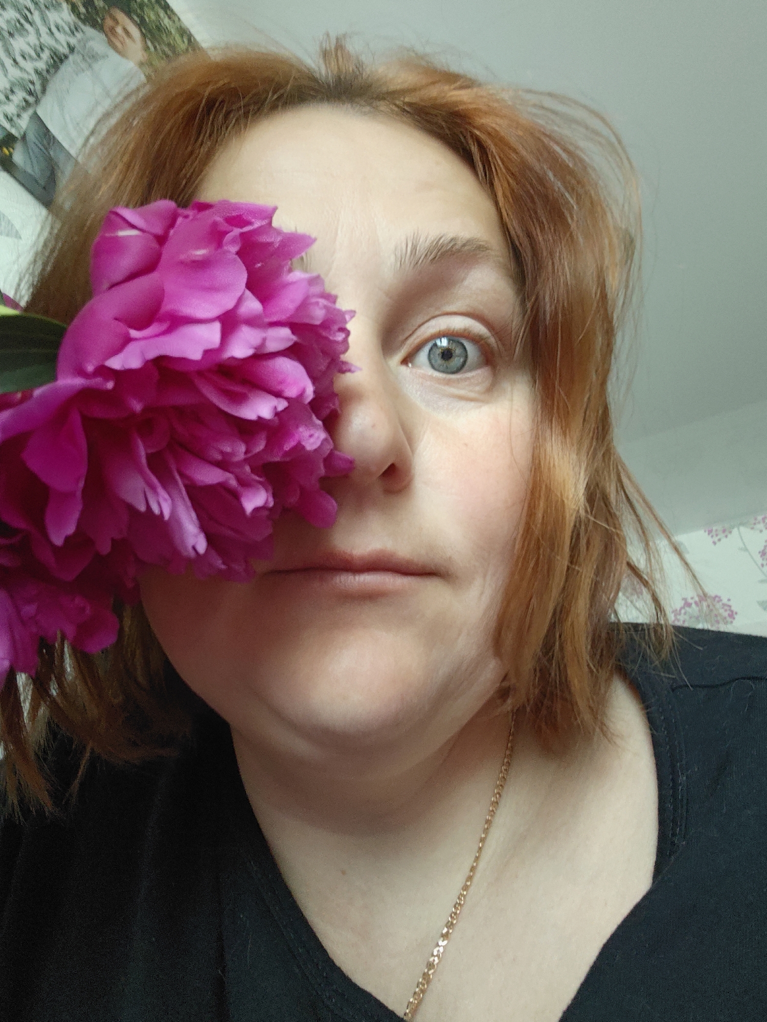 A close-up, low-angle selfie shows a woman with reddish-brown hair and blue eyes holding a large, bright pink peony in front of her face, partially obscuring her left eye and cheek. She is wearing a black t-shirt and a thin gold chain. The background is a light-colored wall with a floral pattern visible on the right. The image appears to be taken indoors. There is no identifiable information that places this image specifically in Markovskii, Russia, or any other city.