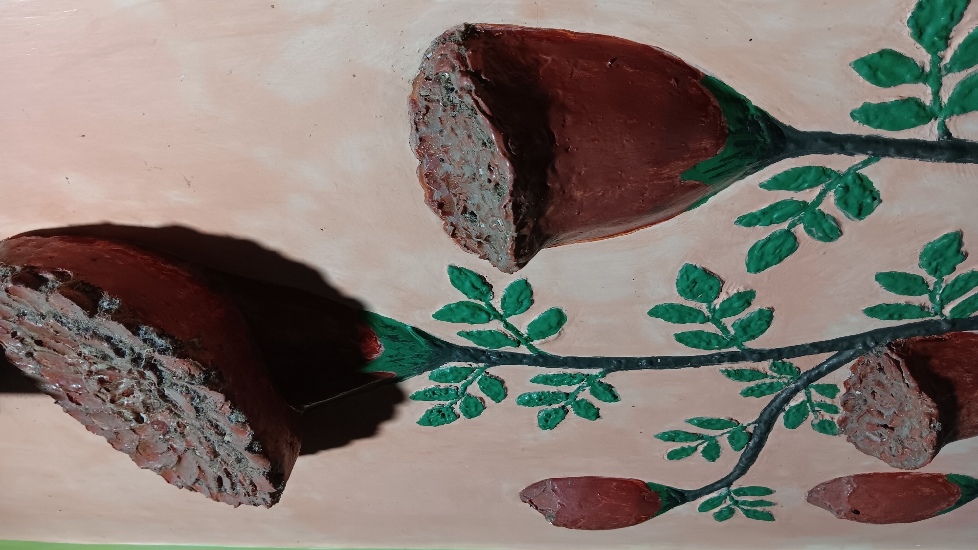 The image depicts a textured wall relief of stylized roses and leaves. The dominant colors are muted pink for the background, reddish-brown for the flowers and buds, and vibrant green for the leaves. Several flowers are shown in various stages of bloom. One large, open flower is at the top left, appearing to be cut in half, revealing a rough, textured interior. Another large open flower is in the middle right, also with a rough, textured interior. To the left, there are two red rosebuds, one slightly lower and to the left of the other. Below the open flower on the right and to the left of the rosebuds, there is a partially visible closed bud with a textured opening. Green leaves are arranged symmetrically on thin, dark stems that extend from the base of the flowers. The lighting suggests the relief is illuminated from the side, casting shadows that accentuate its three-dimensional quality. The overall impression is of a decorative wall art piece. The location in Sonobekel, Indonesia is not evident from the visual information in the image itself.