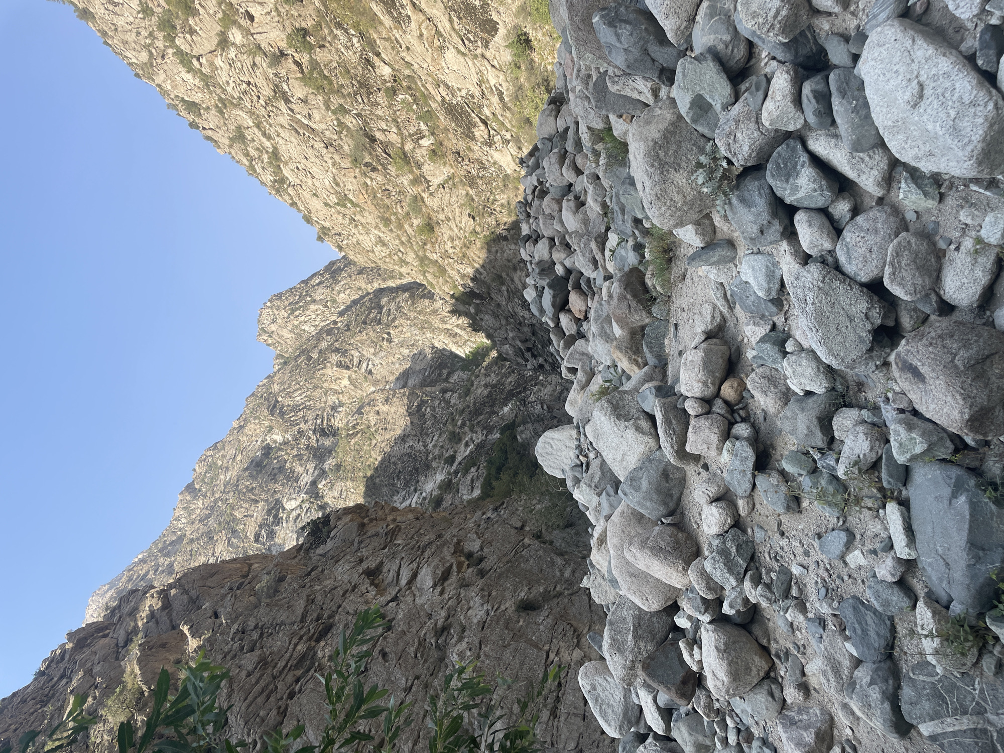 A view looking up a rocky gorge with steep, sun-drenched mountains on either side. The ground in the foreground is covered with a dense collection of rounded grey and tan rocks of varying sizes, interspersed with patches of sparse, dry vegetation. The sides of the gorge are sheer rock faces, showing layers of sediment and erosion. The sky above is clear and blue. This scene is located in the city of Khodzha-Obigarm, Tajikistan.