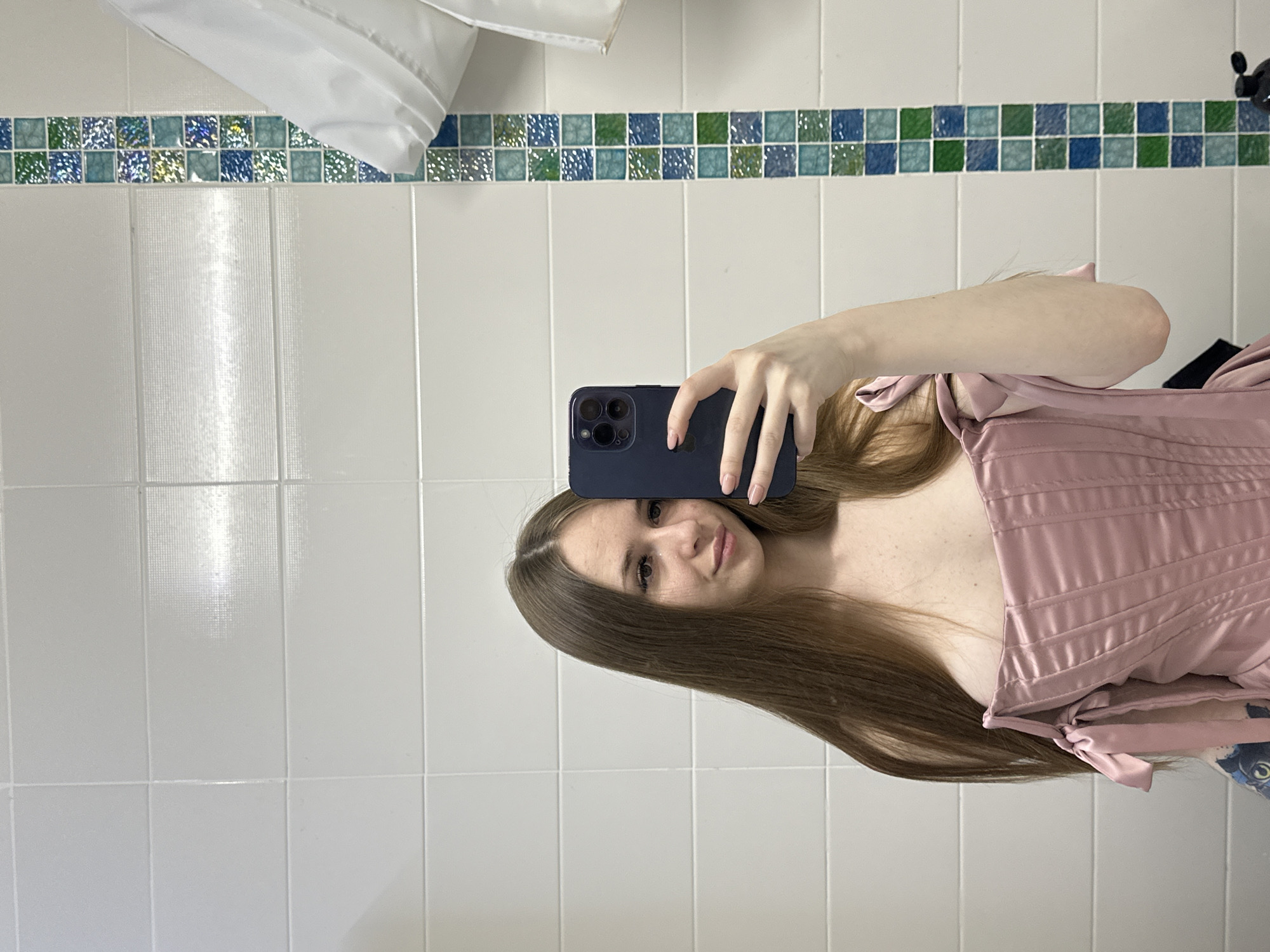 A woman is taking a selfie in a bathroom. The bathroom has white tiled walls with a strip of blue, green, and white mosaic tiles running horizontally near the top. There is a white object, possibly a towel or shower curtain, hanging in the upper left corner. The woman is holding a dark blue smartphone in her right hand, partially obscuring her face. She has light brown hair and is wearing a pink satin top with bow details. The background consists of the bathroom wall and fixtures. The location is unknown.