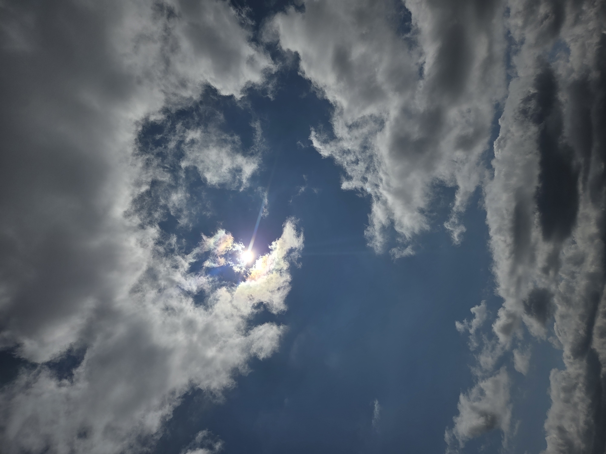 The image depicts a bright sun shining through a gap in the clouds. The sky is blue, with patches of dark and light gray clouds surrounding the sun. The photo was taken in Cherkovitsa, Bulgaria.