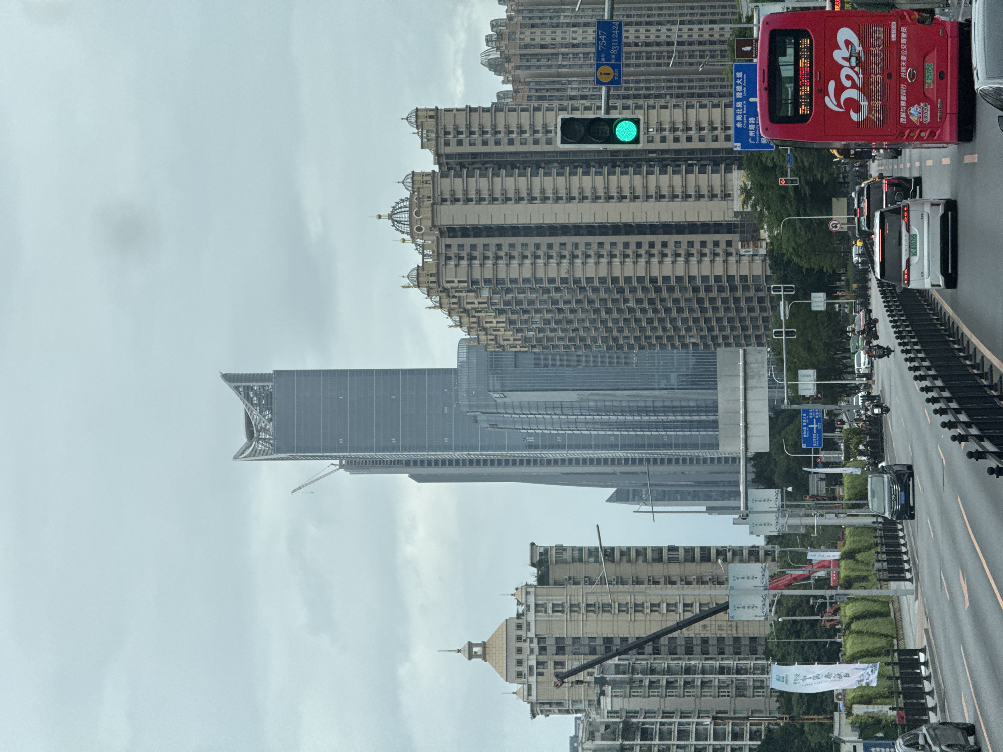 The image depicts an urban scene in Haizhu District, China, likely taken from an elevated position. The foreground shows a multi-lane road with traffic, including a red bus numbered 523 and several cars, motorbikes, and SUVs. Barriers line the edge of the road.
In the middle ground, a row of street signs and greenery separates the road from the buildings. There's a construction crane partially visible amongst the buildings. A tall, modern skyscraper with a distinctive angled top dominates the view, surrounded by other high-rise buildings, some with decorative domes.
The background is filled with more high-rise buildings stretching towards the horizon under an overcast sky. A green traffic light is visible amongst the architecture. There are also street signs that include the words “Chigang North Road” and “Liede Avenue” and “Canton Tower Rd” in Chinese characters.