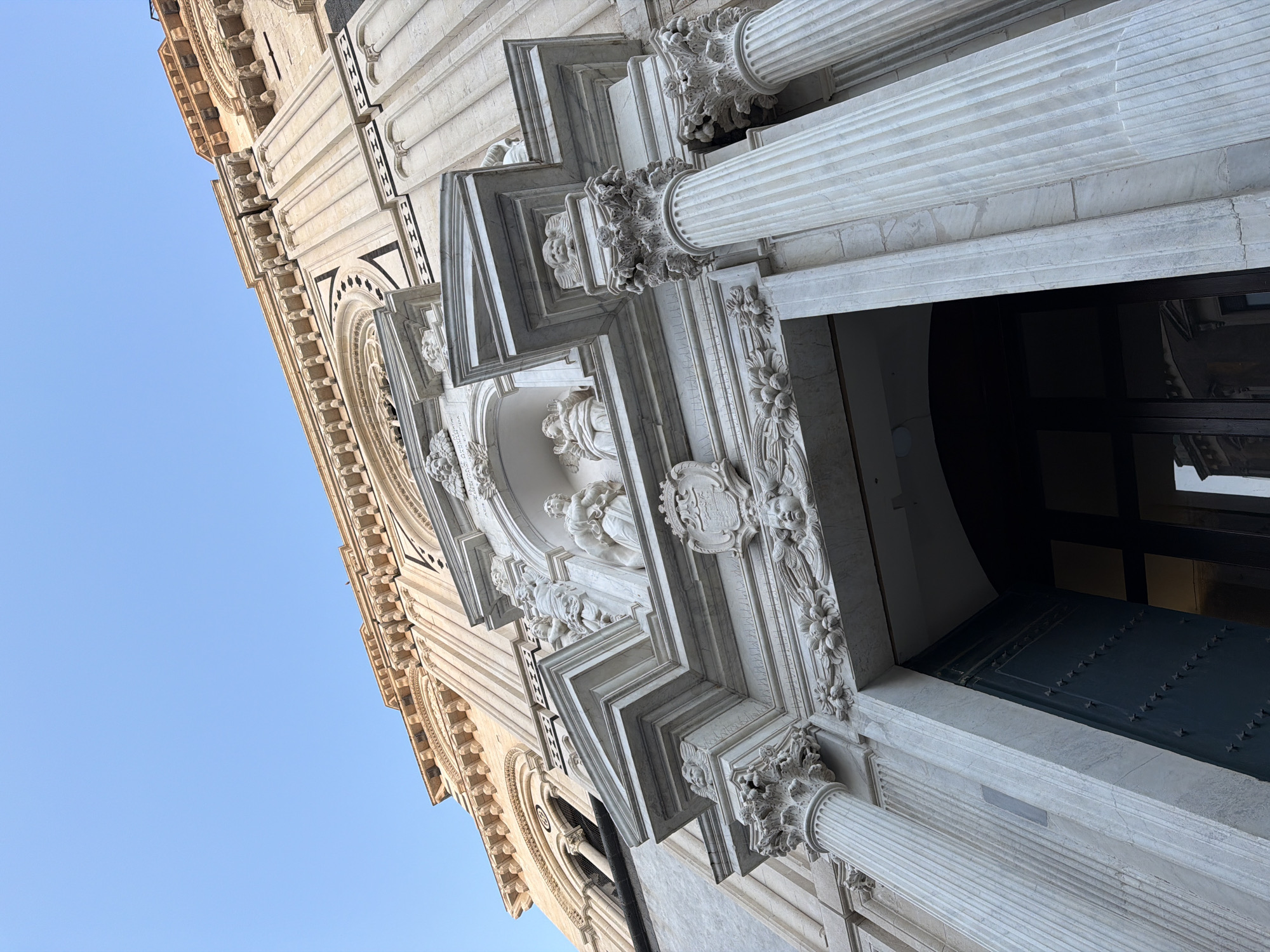 The image shows a low-angle view of the facade of the Basilica di San Sebastiano in Acireale, Italy. The facade is decorated with ornate Baroque sculptures and architectural details, including columns, reliefs, and statues of saints. A doorway is visible in the foreground, with a dark green door. The sky is clear and blue in the background.