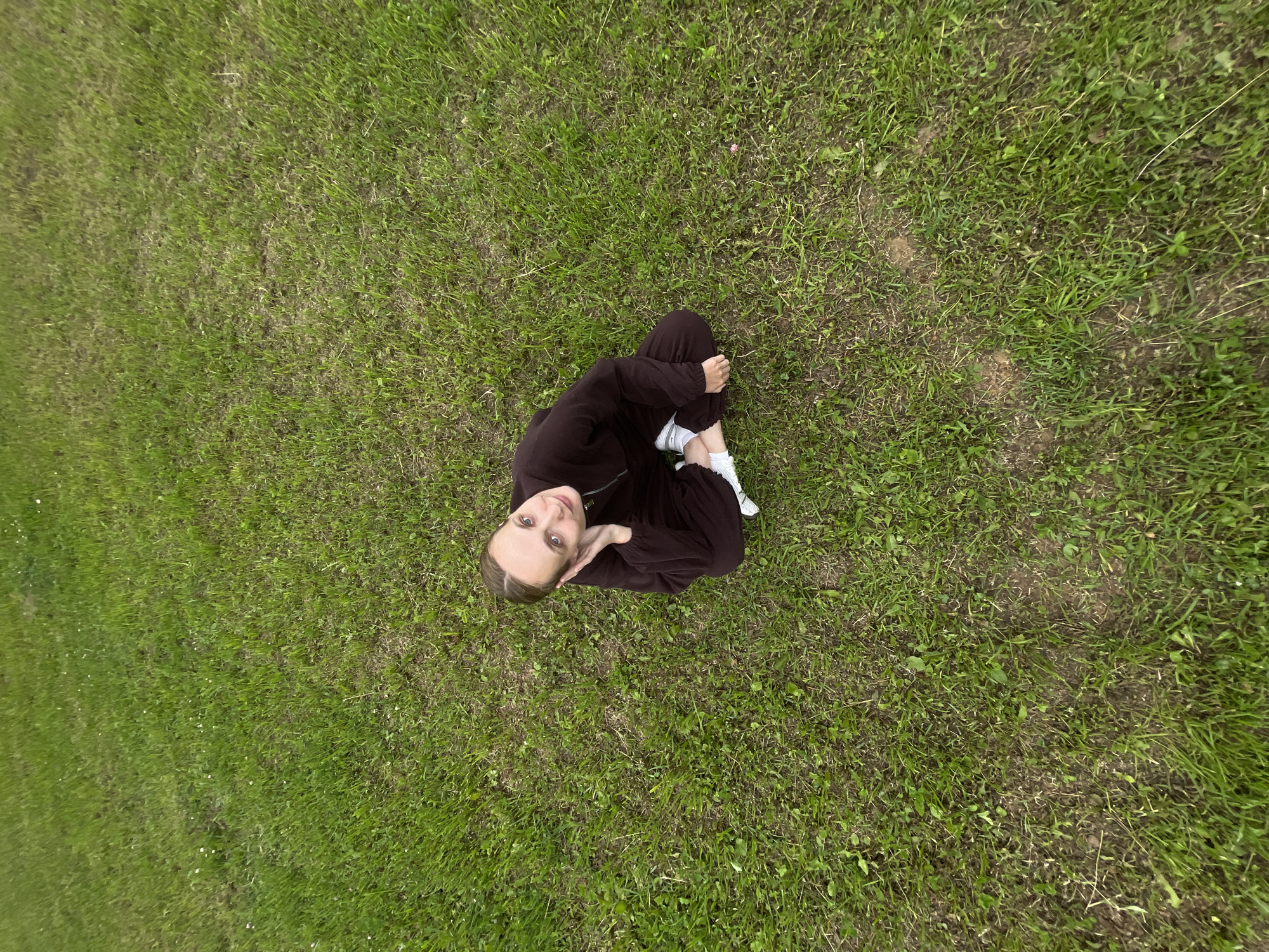 A young woman is sitting on a grassy field in Подшивалово, Russia. She is wearing a dark brown tracksuit and white sneakers, with her legs crossed and her right hand resting on her ear. Her head is tilted back slightly, and she is looking up with a gentle smile. The field is covered in green grass with scattered small white flowers. The perspective is from above, looking down at the woman.