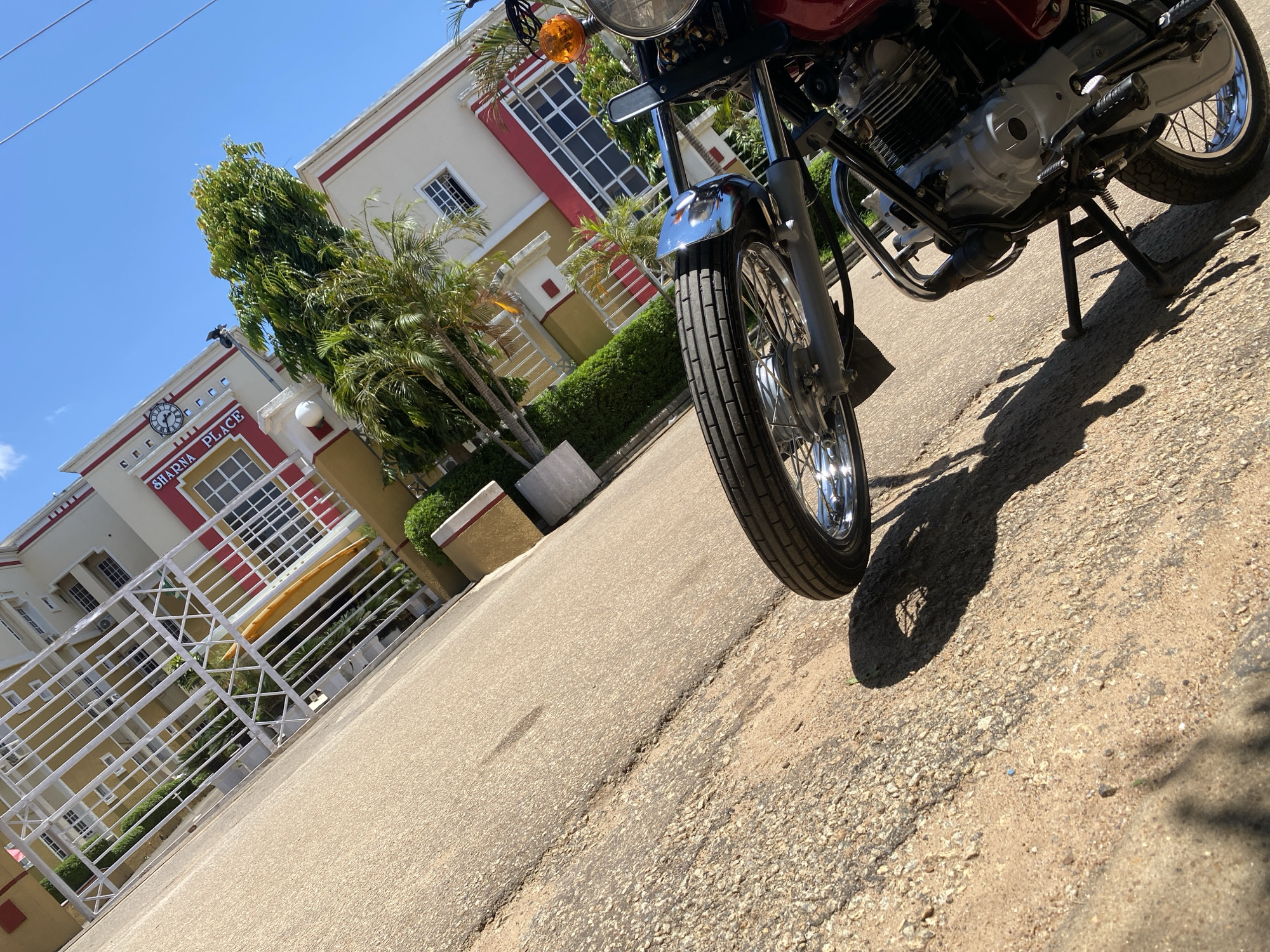A motorcycle is parked on a paved road in front of a building labeled "SHARNA PLACE" in Jos, Nigeria. The building is a multi-story structure with a white and red exterior, featuring multiple windows and a prominent clock. The motorcycle is red and silver, with a visible engine and front tire. The road surface appears worn and cracked, with patches of sand and gravel. Green trees and manicured hedges surround the building. The sky is clear and blue. The image is taken from a low angle, with the motorcycle and road occupying the foreground, and the building in the background.