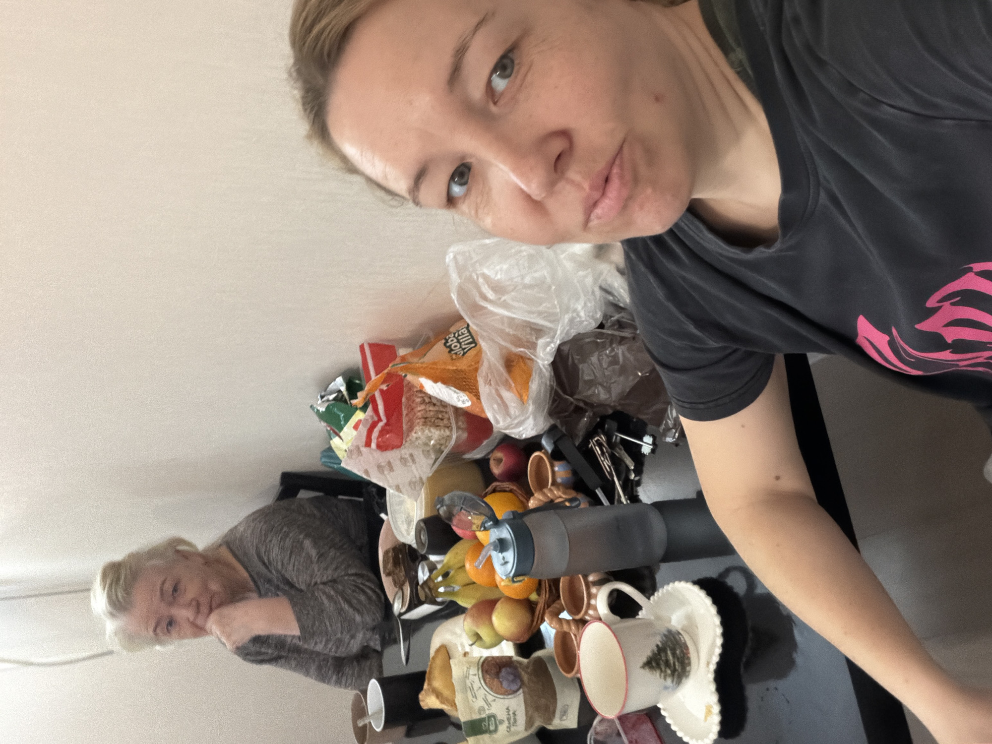 The image depicts an indoor scene in Pervomaysky, Russia, featuring two women seated at a black table laden with various items. In the foreground, a woman in her thirties to forties, with light brown hair and wearing a dark grey t-shirt with a pink design, is capturing a selfie. Her expression is a slight smirk, and her right arm rests on the table. In the background, an older woman, estimated to be sixty to seventy years old, with white or light blonde hair, is seated in a dark chair. She wears a grey long-sleeved top and has her left hand resting on her chin, observing the scene with a thoughtful expression.
The black table between the two women is covered with numerous objects. Directly in front of the foreground woman is a white teacup with a red rim and a Christmas tree design, resting on a white, shell-shaped saucer. To its right, a blue and grey water bottle with a straw lid stands upright. Several small, light brown terracotta-style cups are visible. A wicker basket in the center holds a variety of fresh fruits, including bananas, red apples, oranges, and a green apple. Surrounding the fruit basket are multiple packaged food items, including plastic bags of oranges (one labeled "Global Village"), a package with Cyrillic text, and other unidentified bags and containers. A dark mug and a small, dark plate with a pastry are also present. Keys and other metallic items are scattered on the table. The room's walls are a plain, light, off-white color.