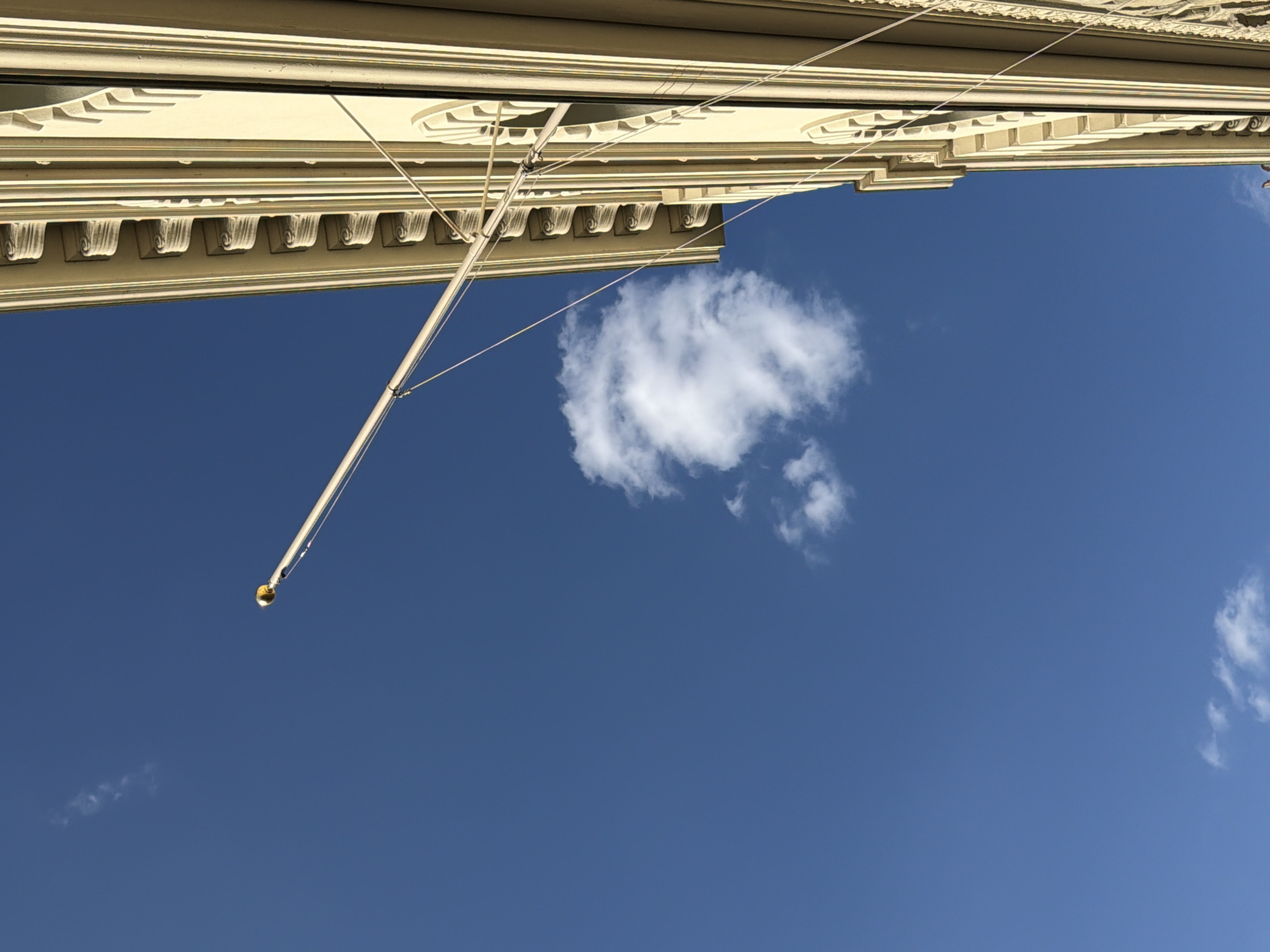 An angled shot captures the upper section of a building with decorative trim against a bright blue sky. A flagpole extends diagonally from the right side of the frame, its tip adorned with a golden finial. A small, puffy white cloud drifts in the sky, positioned below the flagpole. The building's facade features a series of ornate, repetitive elements along its top edge and visible window frames. The image appears to have been taken outdoors, likely on a sunny day. The building's architecture suggests a European style, possibly from the 19th or early 20th century.