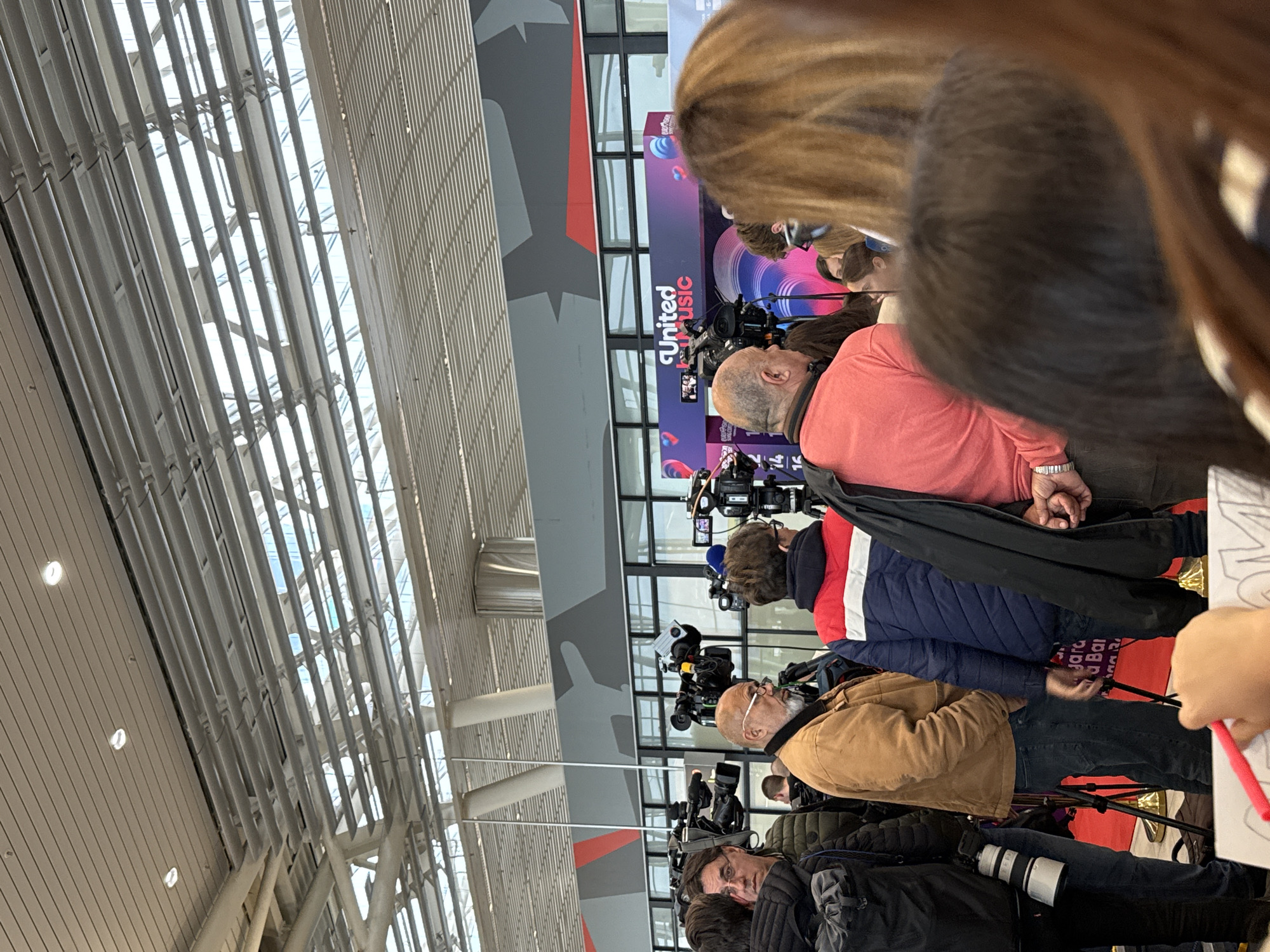 The image shows a crowded scene, likely a press event or media gathering, inside a modern building with a high ceiling featuring exposed structural elements. Numerous cameramen and photographers are present, with cameras and equipment pointed towards a central point of interest. A backdrop is visible with the text "United Music," indicating a music-related event. People are standing close together, with some holding microphones. The location appears to be an airport due to the architecture and signs. The specific airport in Sofia, Bulgaria, cannot be determined from the image alone.