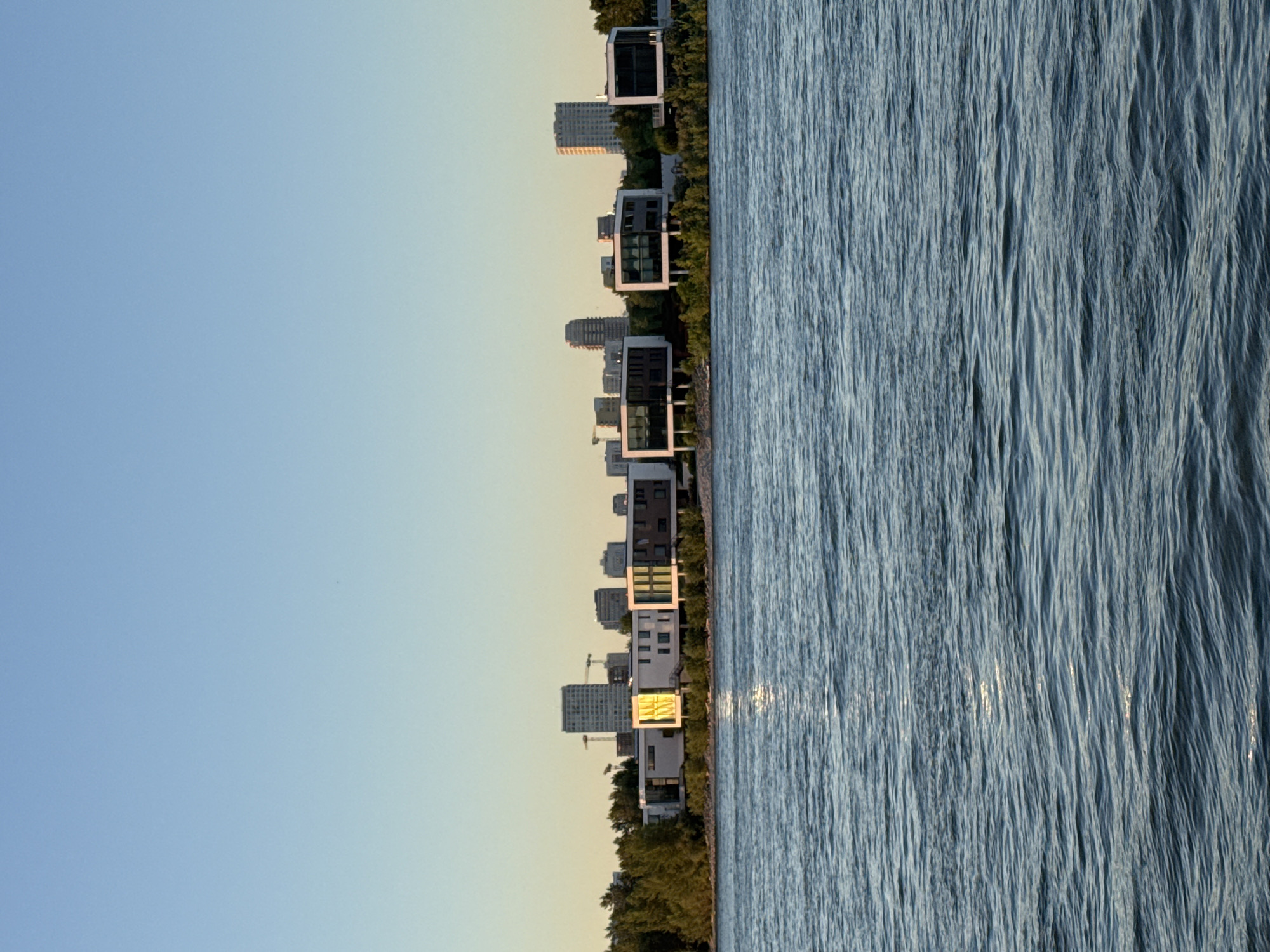 The image shows a wide body of water in the foreground, with gentle ripples reflecting the clear blue sky and the warm glow of the setting sun. In the middle ground, a line of modern, multi-story buildings with large windows and white facades runs along the waterfront. Some of these buildings feature prominent yellow accents. Behind these structures, the silhouette of a city skyline is visible, with several taller buildings and construction cranes dotting the horizon. Lush green trees and bushes frame the waterfront buildings. The overall impression is of a serene, contemporary urban landscape at dusk, situated along a river or large body of water. This scene is consistent with the area along the Ob River in Novosibirsk, Russia, where modern residential and commercial developments can be found.