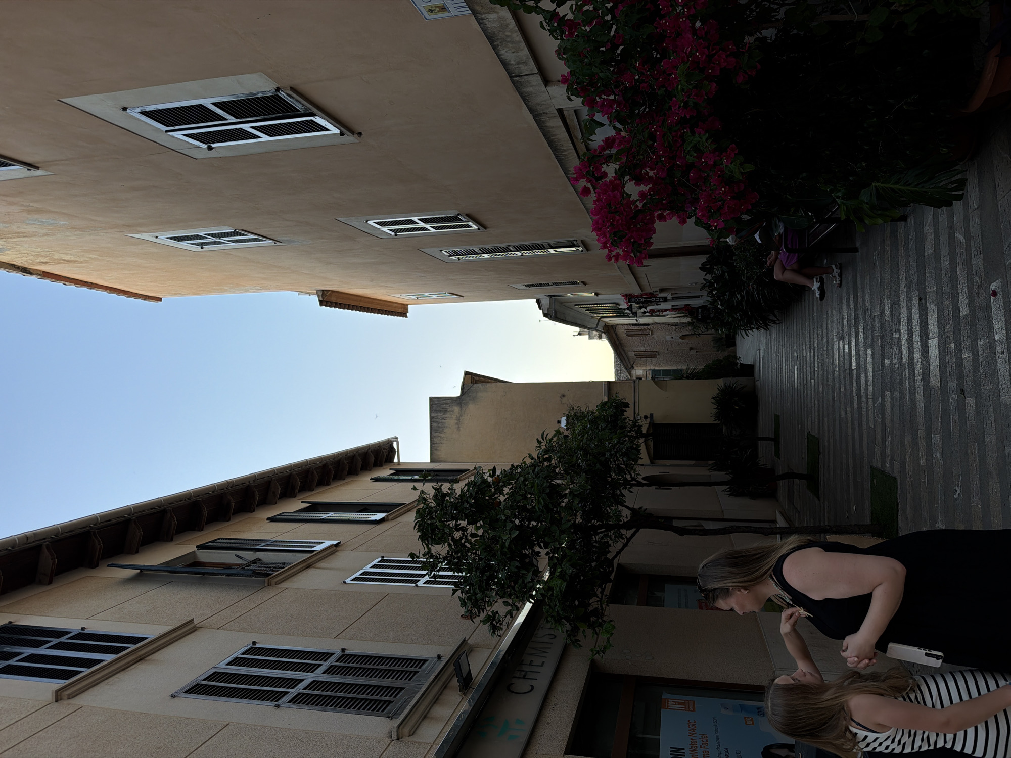 The image depicts a street scene in Alcúdia, Spain, with buildings lining a pedestrian walkway. The buildings are light-colored with dark shutters on the windows. A bougainvillea plant with pink flowers cascades from a building on the right side of the image. People are present in the lower right corner of the image, including two women. The image is oriented so that the sky is on the left side, and the building facades are oriented vertically.