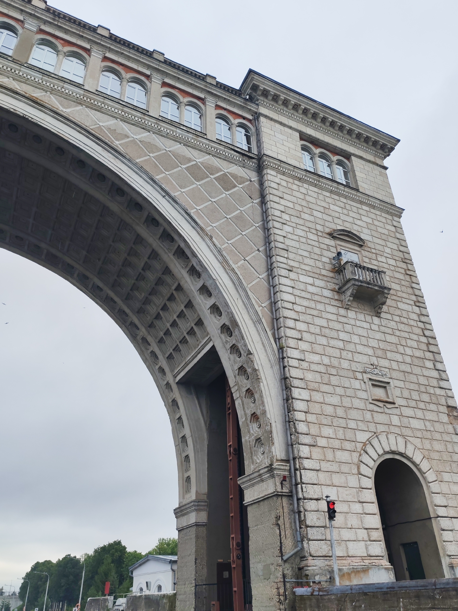 The image shows a large, stone archway with a tower on the right. The archway has decorative patterns and is supported by massive stone blocks. The tower is made of lighter colored stone blocks and has several windows. There is a small balcony on the tower. A traffic light with a red signal is visible in front of the tower. The sky is overcast. Trees and some buildings are visible in the background. The overall scene suggests an architectural structure of historical significance, possibly a bridge or part of a canal system. The location is Faliukovo, Russia.