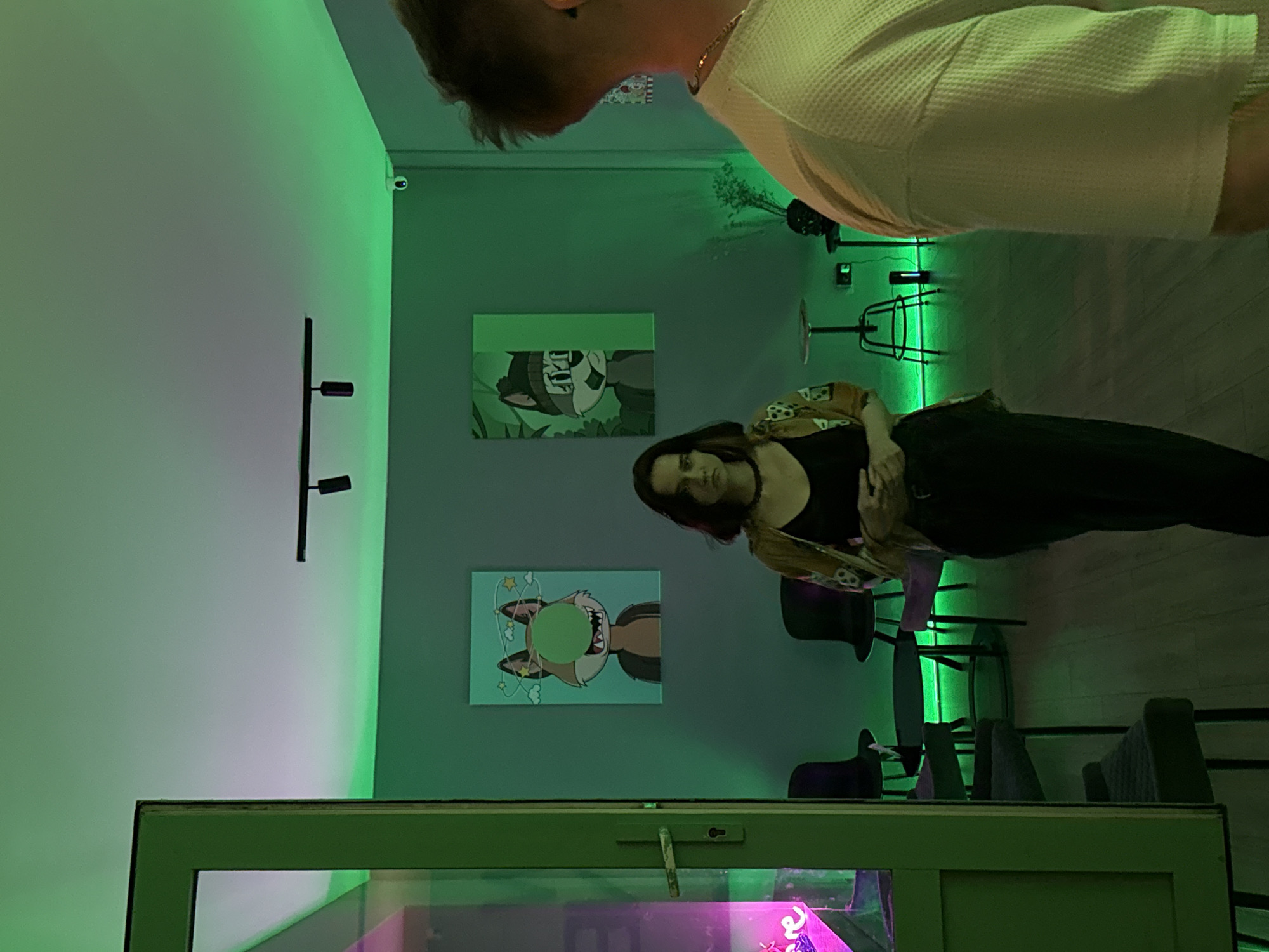 The image depicts an interior space, illuminated by green and purple lighting. Two people are present, one partially visible from above, wearing a textured white shirt and a gold chain, and the other standing in the middle ground wearing a black top, black pants, and a patterned jacket. On the wall are two framed cartoon-like images. There are chairs and bar stools along the right side. In the foreground is a window or display case illuminated with bright pink lighting. The location is likely in Belgrade, Serbia, but there is no specific visual information to confirm this.