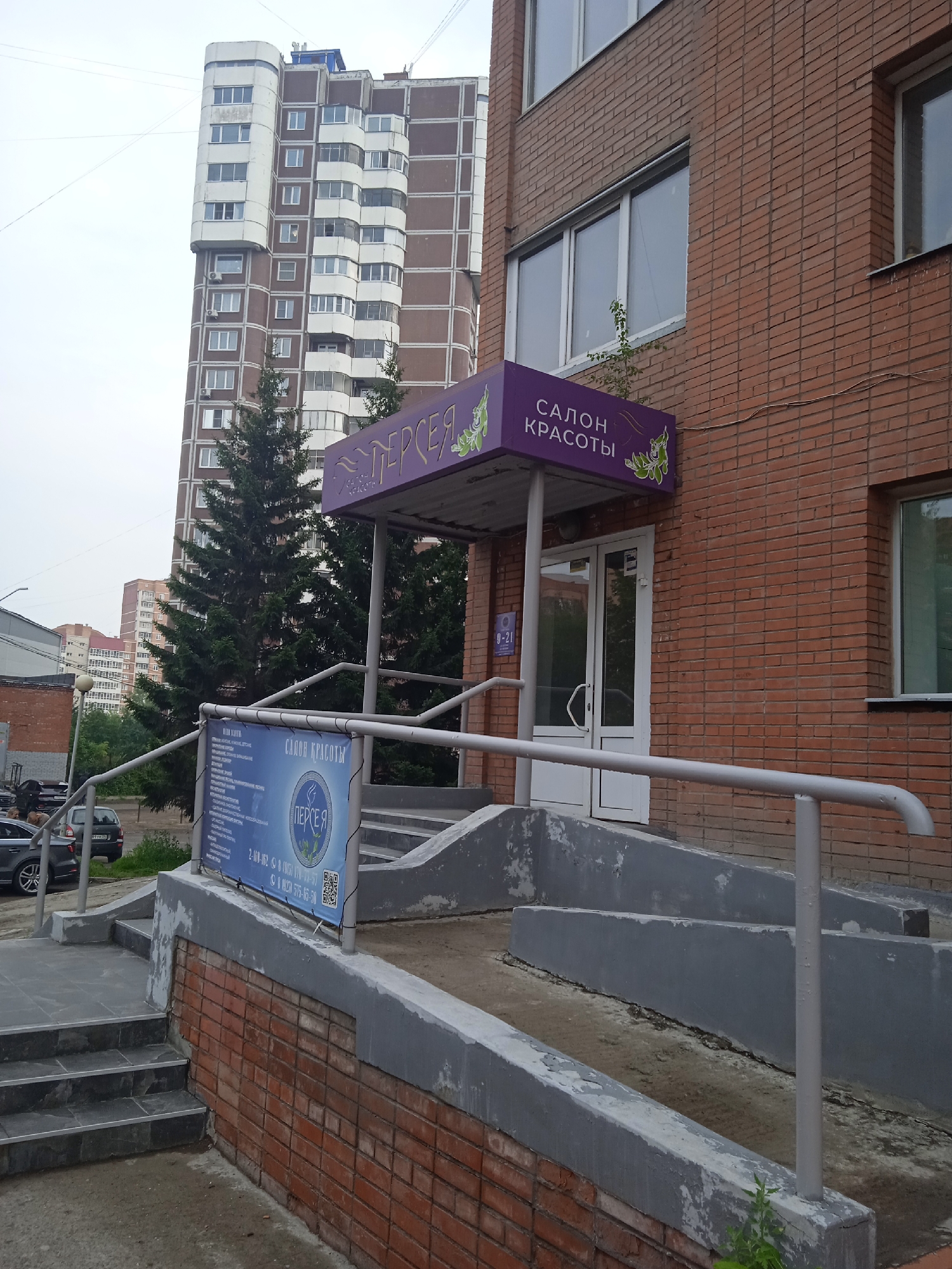 The image shows the entrance to a beauty salon named "ПЕРСЕЯ" (Perseya) located in Krasnoyarsk, Russia. The salon has a purple awning above the entrance with the words "САЛОН КРАСОТЫ" (Beauty Salon) written in white letters. Below the awning, there is a double glass door. To the left of the entrance, there is a blue banner with text and a QR code, which is likely an advertisement for the salon's services. A ramp with a metal railing leads up to the entrance, and to the left of the ramp, there are stairs. The building is made of red brick, and there are large windows on the upper floors. In the background, a tall, modern apartment building with a brown and white facade is visible, along with some evergreen trees. Several cars are parked on the street in front of the building. The sky is overcast.