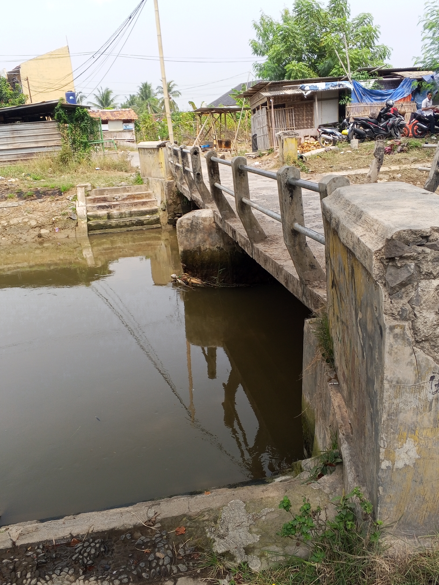 A concrete bridge spans a murky body of water. On the left bank, stone steps lead up to the bridge. The right bank is overgrown with grass and vegetation. In the background, several dilapidated structures, including a building with a blue tarp roof and what appears to be a motorcycle repair shop, are visible. A cluster of motorcycles is parked near the buildings. A lone figure stands near the buildings, and another sits on a motorcycle. The sky is overcast. The image appears to be taken from the bank of the water.