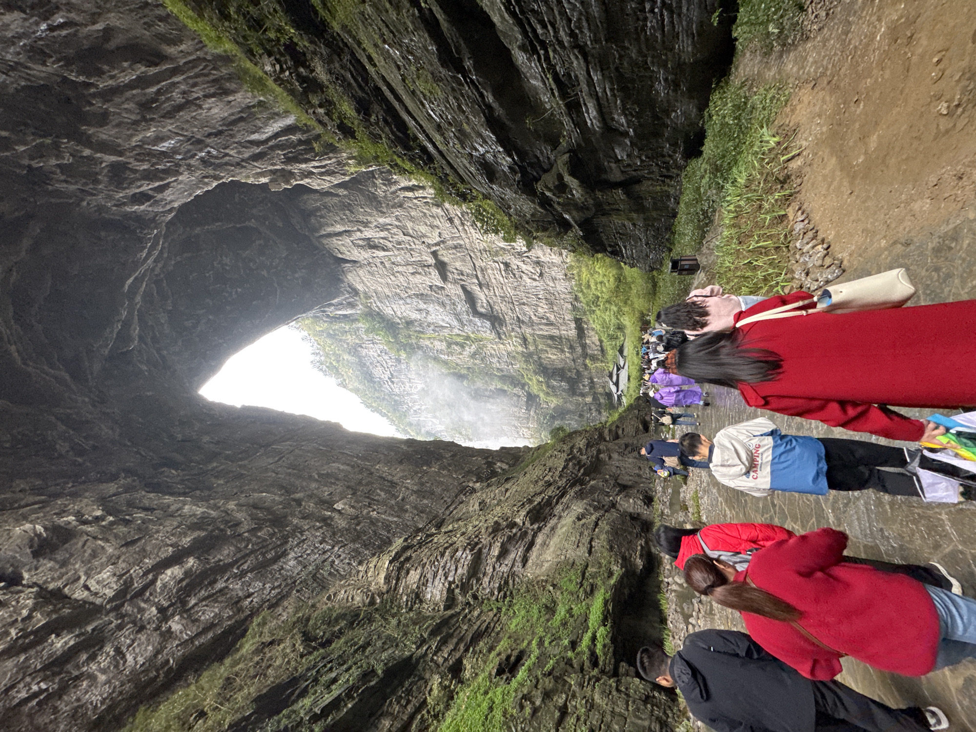 A view from the bottom of a cave in Wulong District, China, showing several people walking away from a light source at the opposite end. The cave walls are tall and rocky, with patches of green vegetation. Most people are wearing casual clothing, with some wearing red jackets and one in a purple raincoat. The ground appears wet, possibly a shallow stream or standing water.