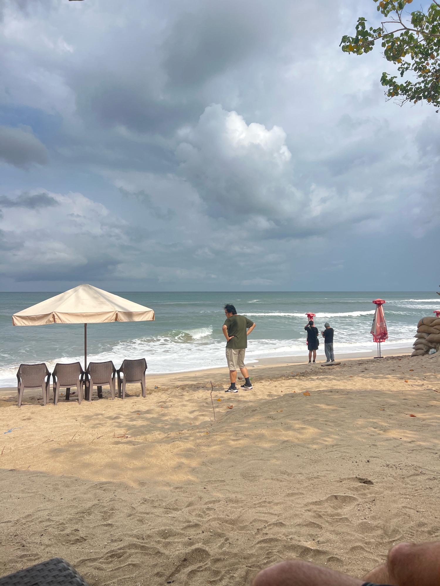 An overcast day on Kuta Beach, Bali, Indonesia, features a sandy shoreline with the ocean waves rolling in. In the foreground, a woven chair is partially visible at the bottom of the frame. Further back, four empty plastic chairs sit beneath a large, cream-colored beach umbrella. To the right, two red beach umbrellas are set up, one with a red canopy and the other with a red and white striped canopy. Several sandbags are stacked next to one of the umbrellas. Three people are on the beach: one man in a green t-shirt and beige shorts stands with his hand on his hip, facing away from the water. Two other individuals, one in a black shirt and the other in a grey shirt, stand closer to the water's edge, appearing to interact with their phones. The sky is filled with heavy, grey clouds, suggesting the possibility of rain.