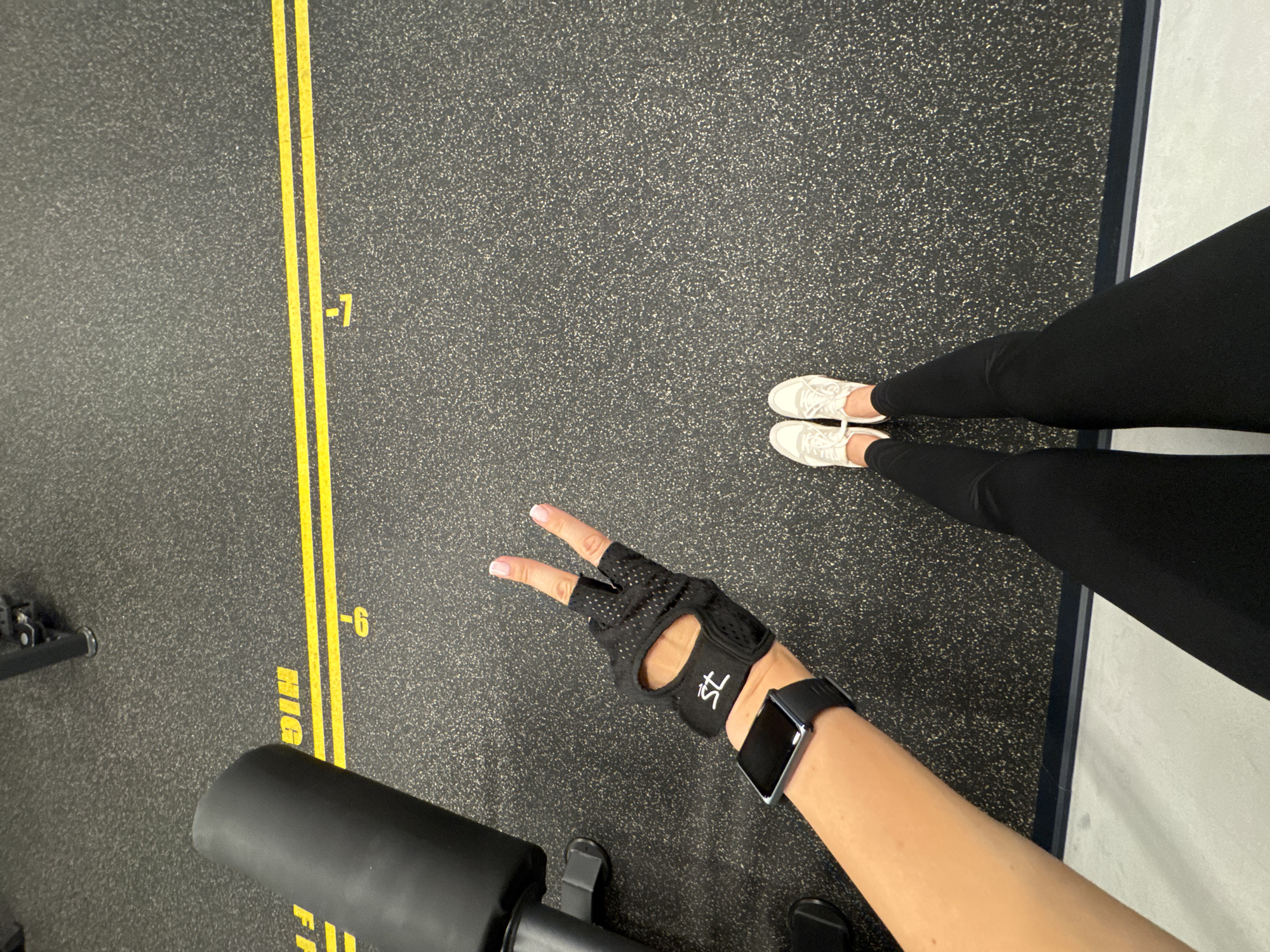 The image shows a first-person view of a person in a gym, likely during a workout session. The scene is set on a dark, speckled rubber gym floor with yellow markings. On the floor, there are two parallel yellow lines with markings indicating numerical positions: "-7" and "-6" are visible. Part of a black gym bench is in the foreground. The person is wearing black leggings and white sneakers. Their right arm is extended, wearing a black fitness glove with mesh and a white logo on it. The hand is making a peace sign with two fingers extended. A black smartwatch is worn on the wrist. The overall impression is of an active and healthy lifestyle, with the gym setting and fitness attire suggesting a workout is in progress or has just concluded. The lighting appears to be artificial, common for indoor gym environments. No specific time of day or weather conditions can be determined from the image. No identifiable text is visible beyond the numerical markings on the floor and the logo on the glove. The location context of Sochi, Russia, is not directly evident from the visual cues within the image itself.
