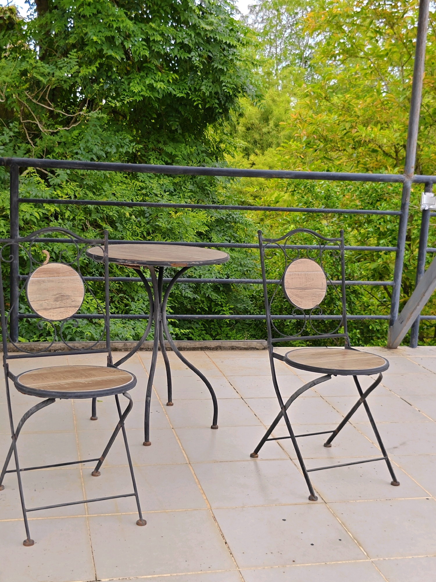 This is a medium shot, eye-level, outdoor photograph of a wrought iron patio set on a tiled balcony. The set consists of one round table and two chairs, all with wooden slatted tops and seats, and decorative wooden circles on the chair backs. The furniture is dark metal with some rust. The balcony is paved with square beige tiles. In the background, a black metal railing runs horizontally, behind which is lush green foliage from trees, with some yellowing leaves mixed in. The railing and foliage suggest the location is elevated, possibly a second-story balcony or a terrace. The setting appears to be in Mu Si, Thailand, given the dense tropical greenery and the style of the outdoor furniture often found in such locations. There are no people in the image.