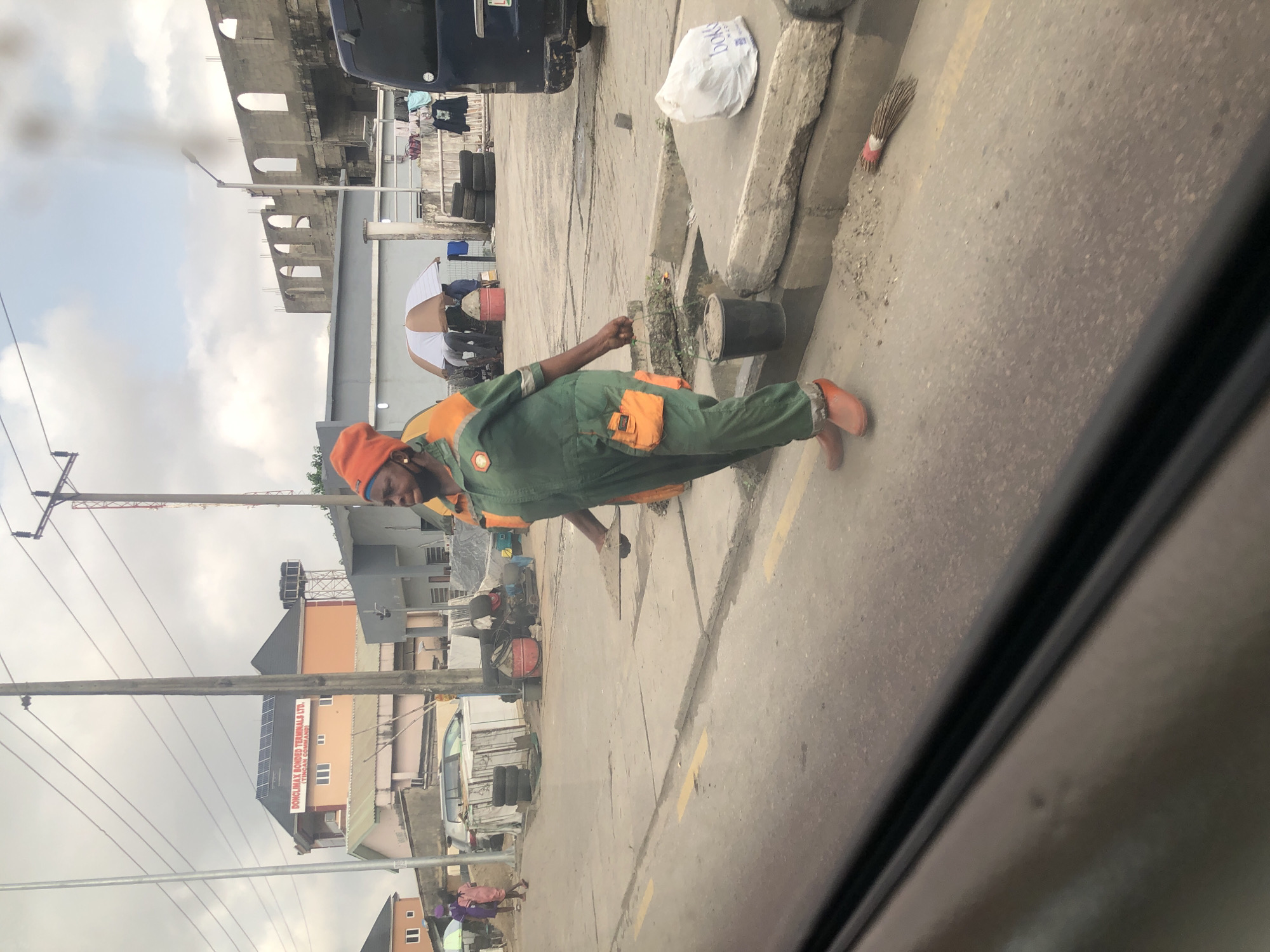 In Agani, Nigeria, a street scene unfolds with a focus on a sanitation worker. The individual, dressed in a green jumpsuit with orange accents and an orange beanie, is captured mid-stride. They are carrying a small bucket in their right hand, which appears to contain dirt. The worker's footwear consists of bright orange boots. Behind them, another sanitation worker in a similar uniform is partially visible. The environment suggests a commercial or industrial area, with buildings under construction in the background, including structures with arched windows. Various objects are scattered on the ground, such as tires, construction materials, and a white plastic bag. A broom lies on the pavement near the curb. Power lines crisscross the sky, which is overcast. The road is paved and marked with yellow lines.